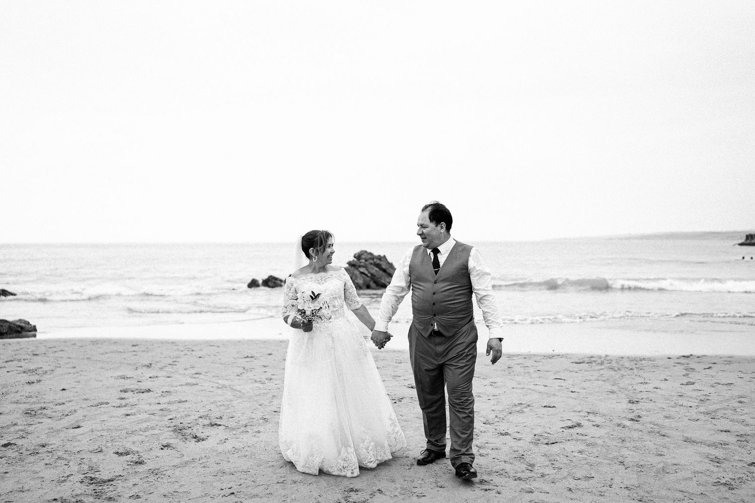 A couple walks hand in hand on a beach during their black and white wedding photoshoot.