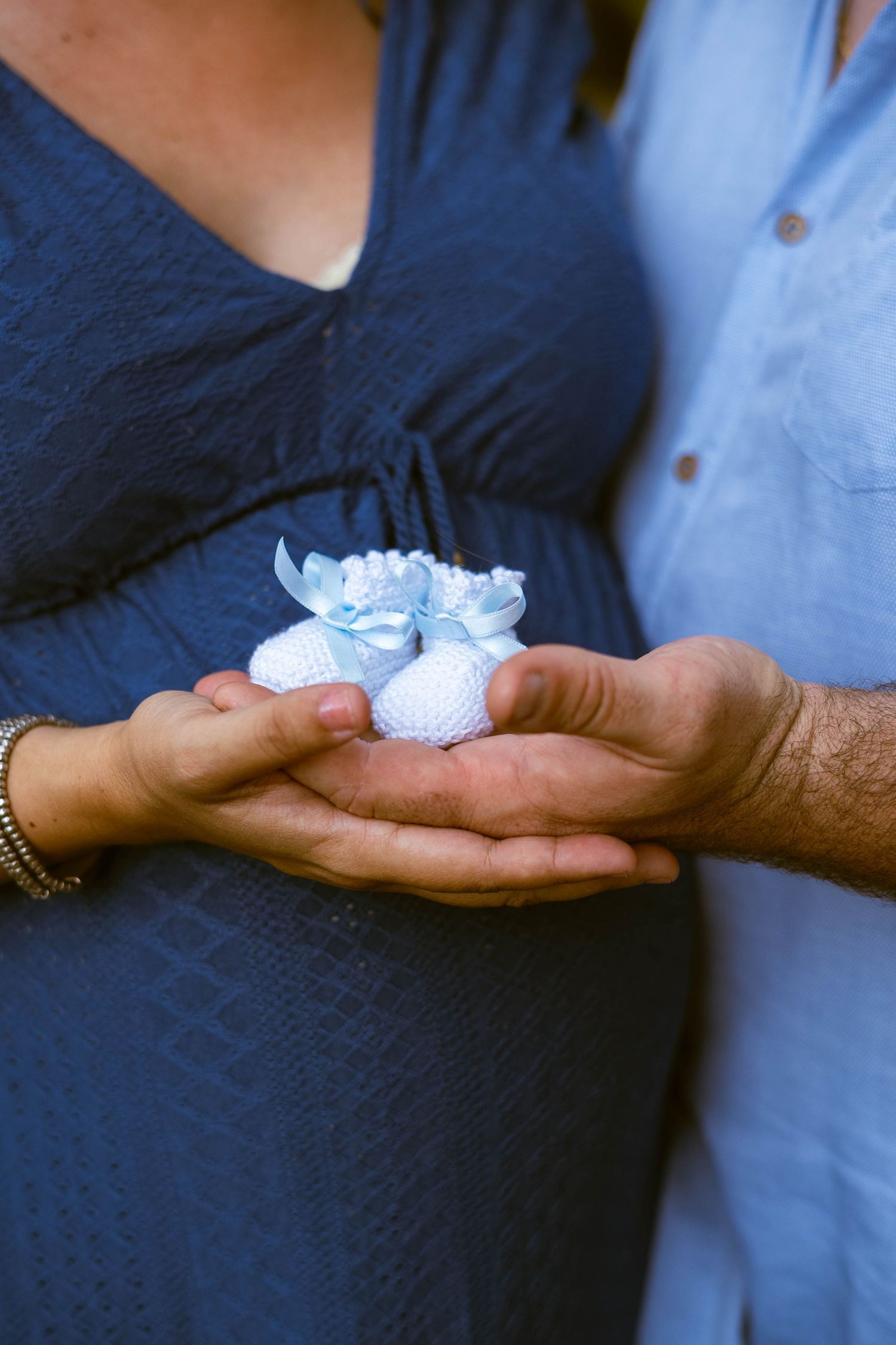 Hands holding delicate blue baby booties against a navy and light blue clothing background.
