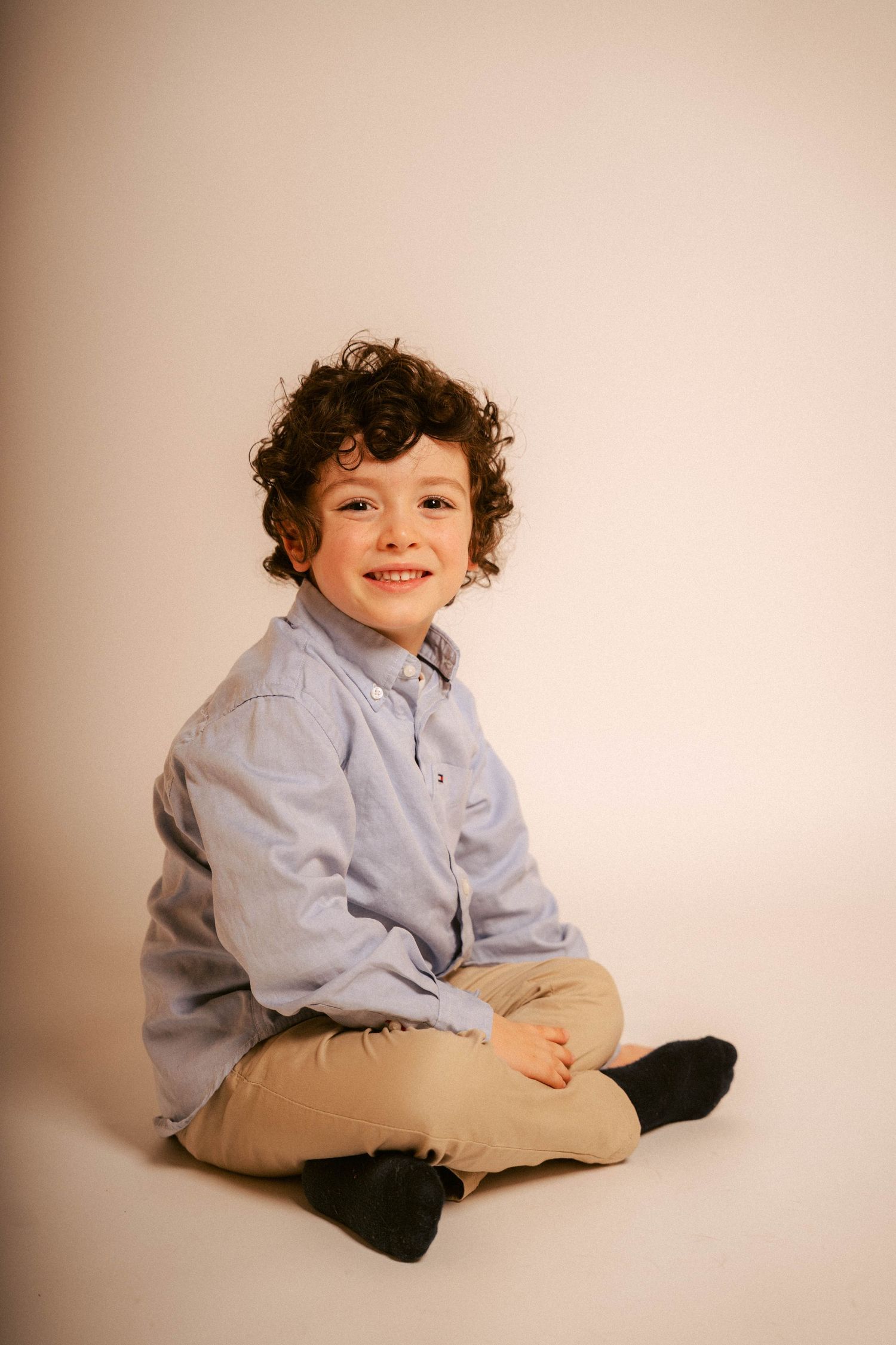 Young child in light blue button-down shirt and khaki pants sits and poses on beige backdrop in studio setting.