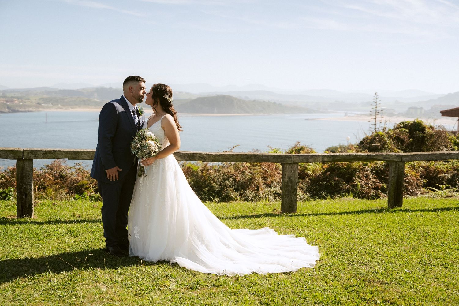 A couple embraces on a grassy hilltop overlooking a scenic coastal bay on their wedding day.