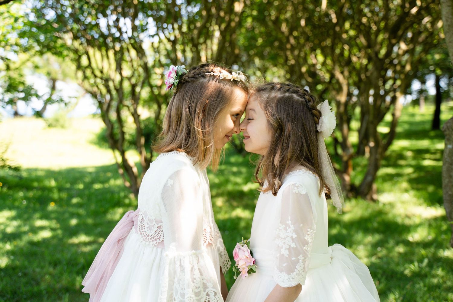 Two children in white dresses hold hands while walking through a garden path surrounded by greenery.