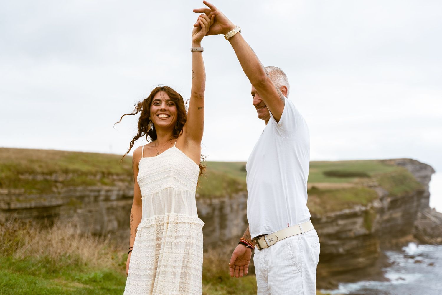 A couple in white outfits dance together on a grassy cliff overlooking the ocean.
