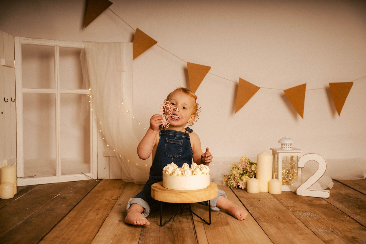 A baby in a blue outfit sits on a wooden floor during a first birthday celebration with bunting decorations.