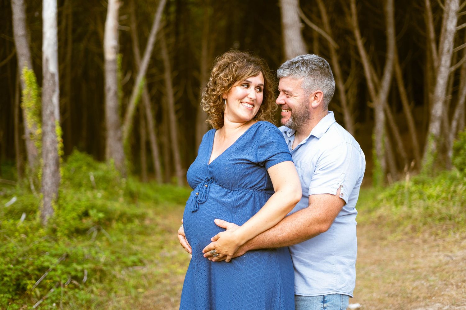 Expectant couple embracing in a wooded setting during sunset.