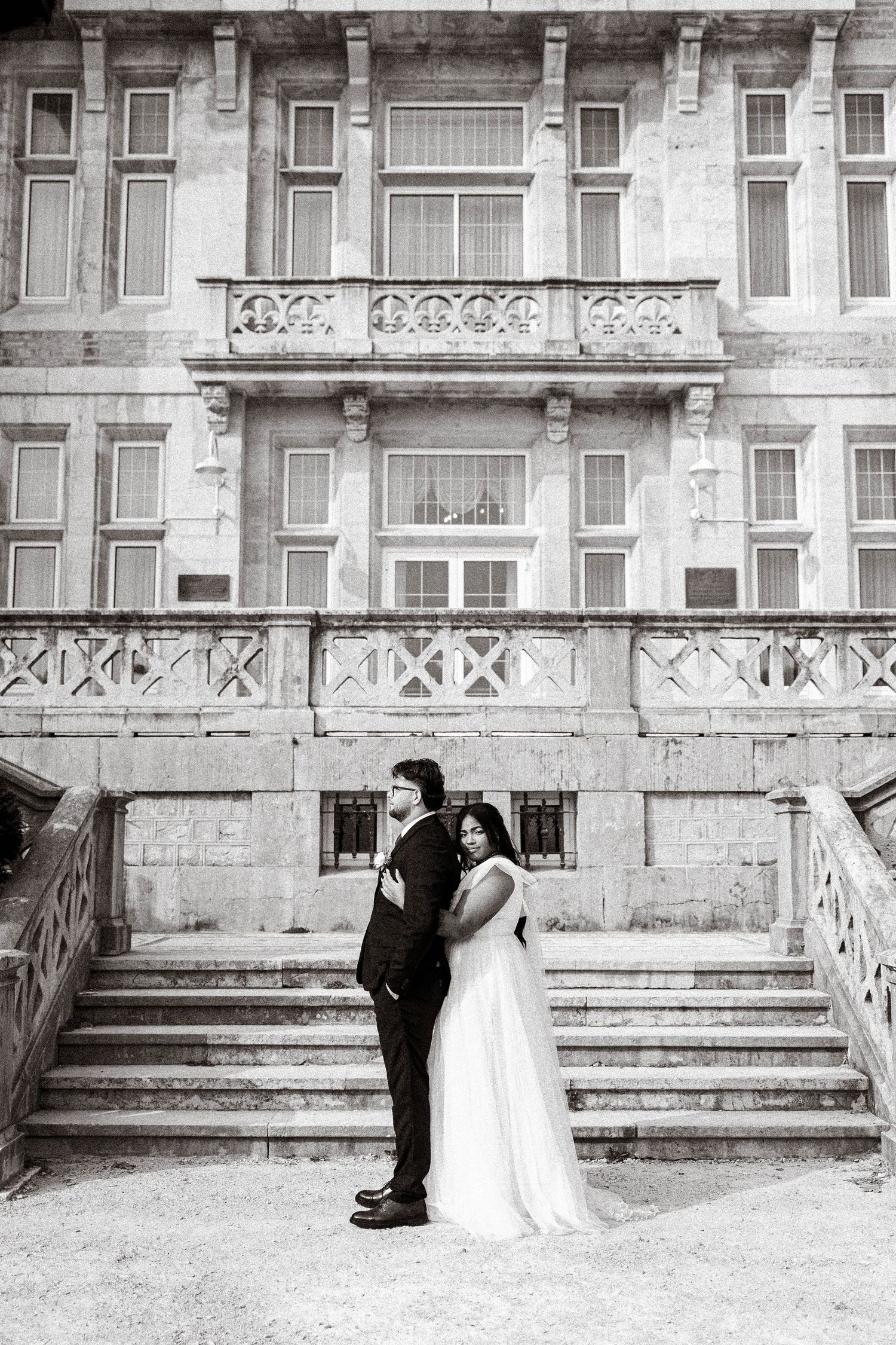 A bride and groom pose on grand stone steps in front of a historic building in black and white.