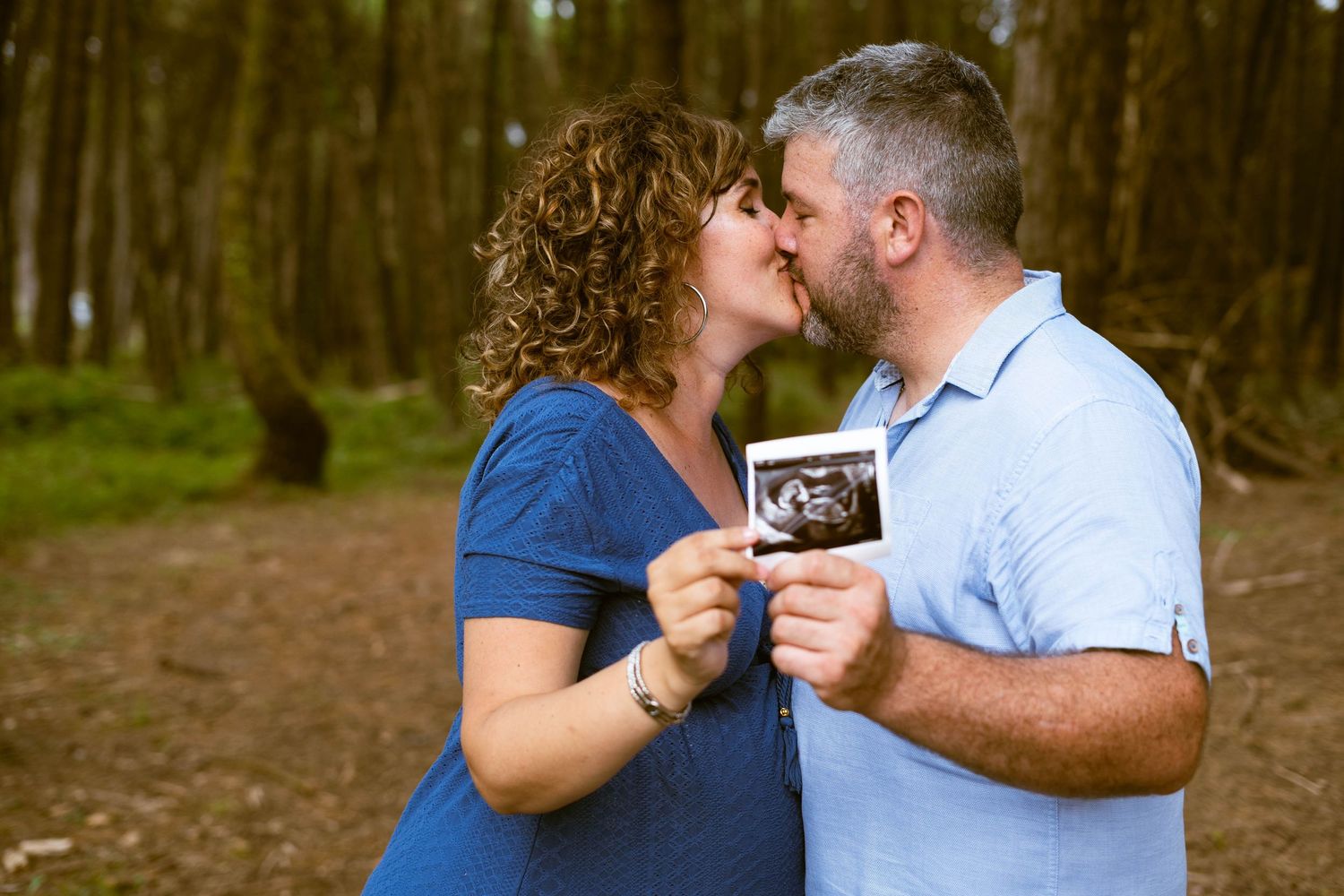 Couple sharing a kiss while holding an ultrasound photo in a forest setting.
