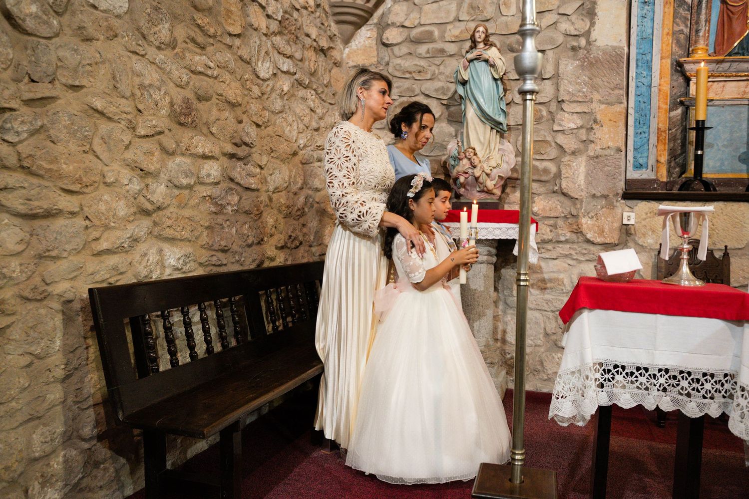 Children in white formal wear stand near altar inside historic stone church with red accents.