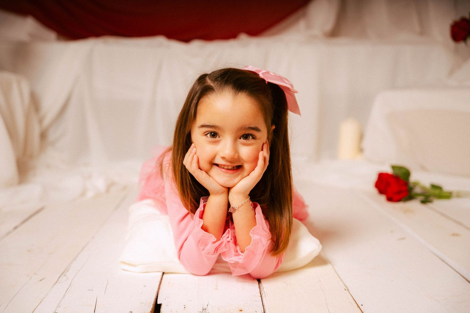Young child in pink dress poses playfully on white floor with red rose in the background.
