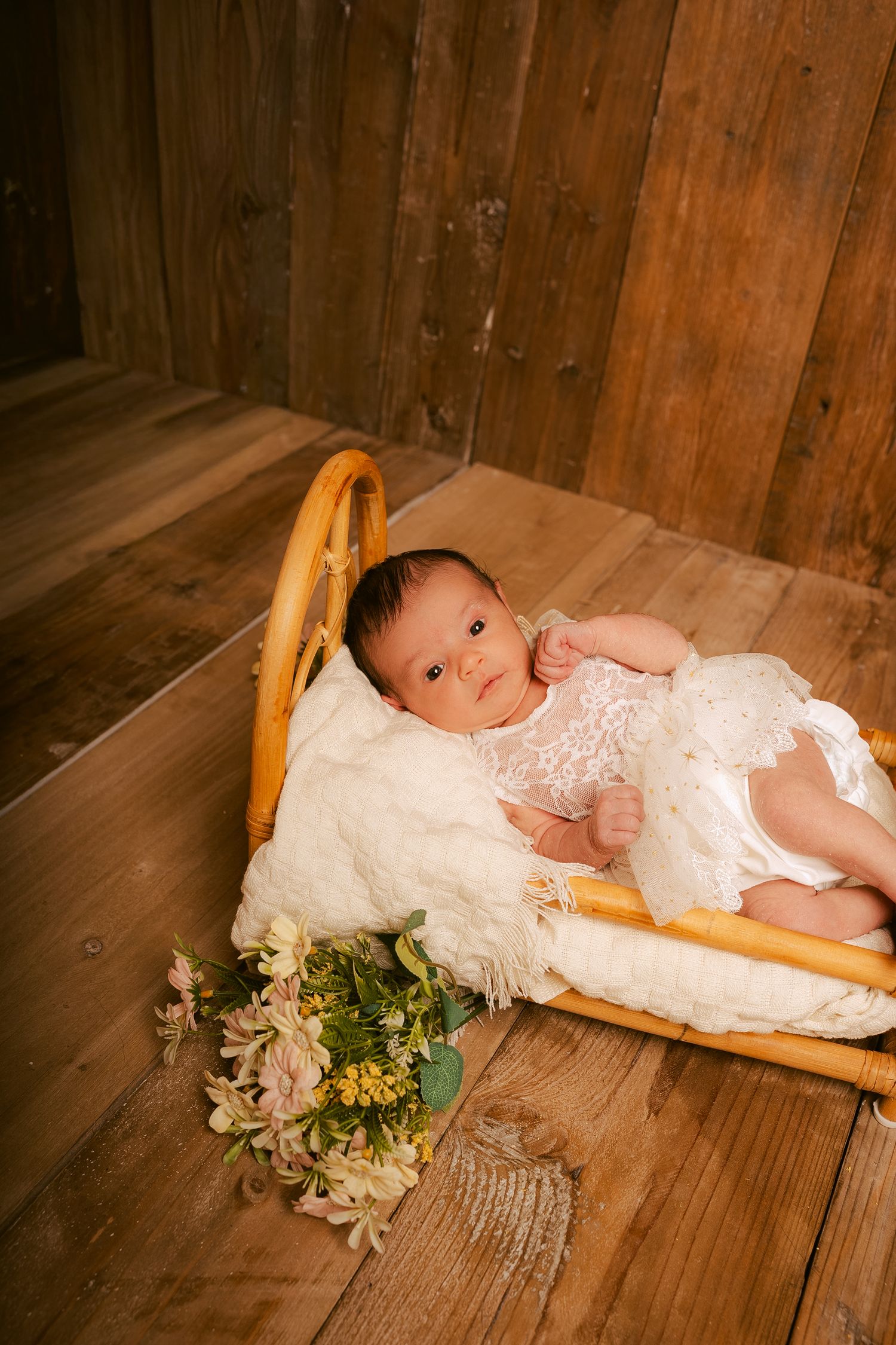A baby in white lace dress lies in a wooden basket with flowers on rustic hardwood floor.
