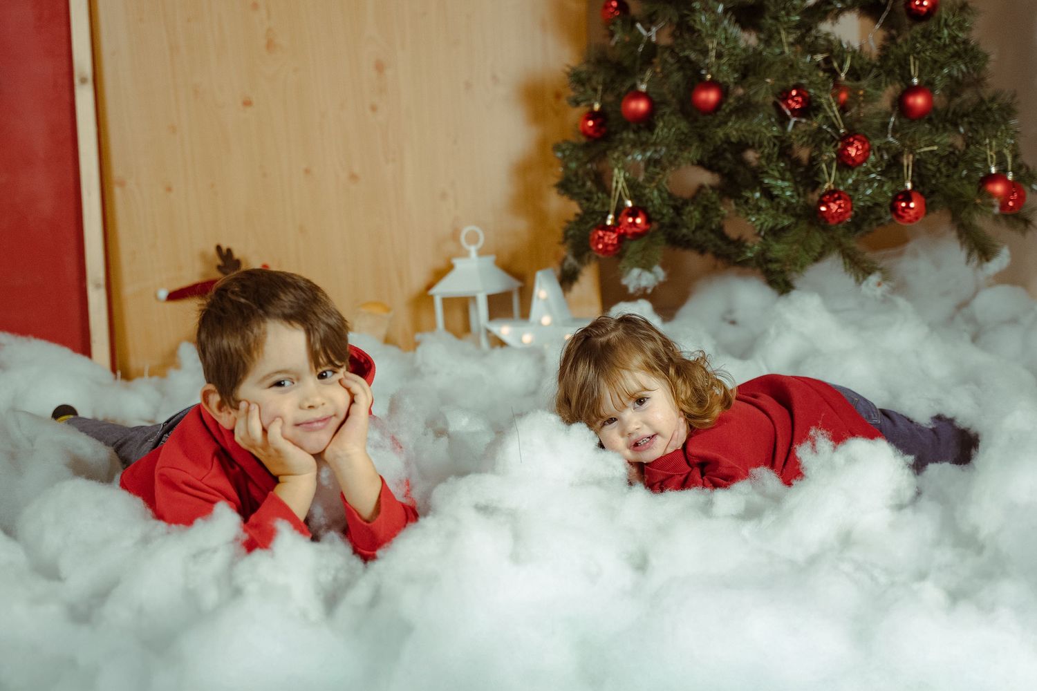 Two children in red outfits lay in fluffy white cotton snow in front of a decorated Christmas tree.
