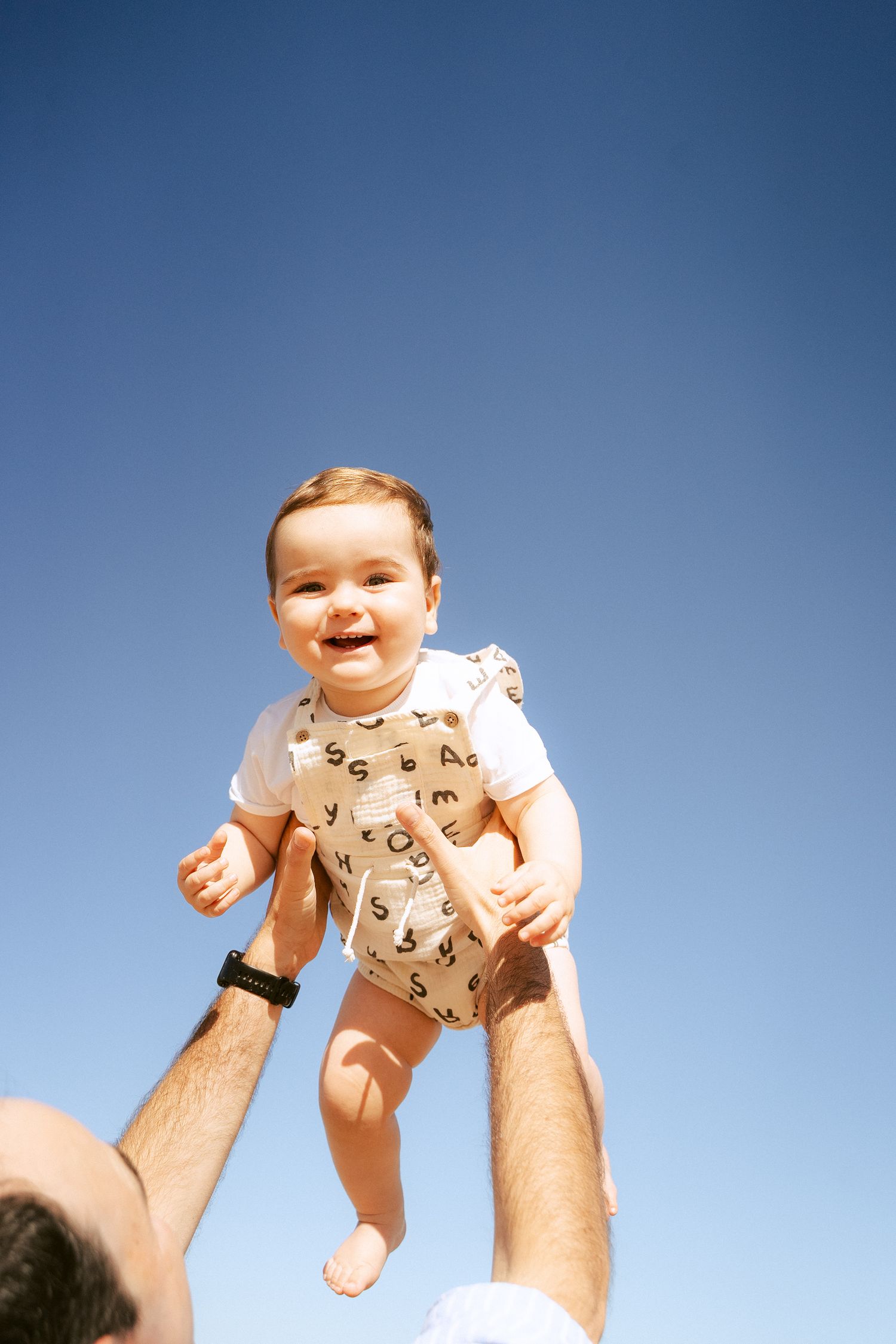 Baby in onesie being lifted against blue sky background.