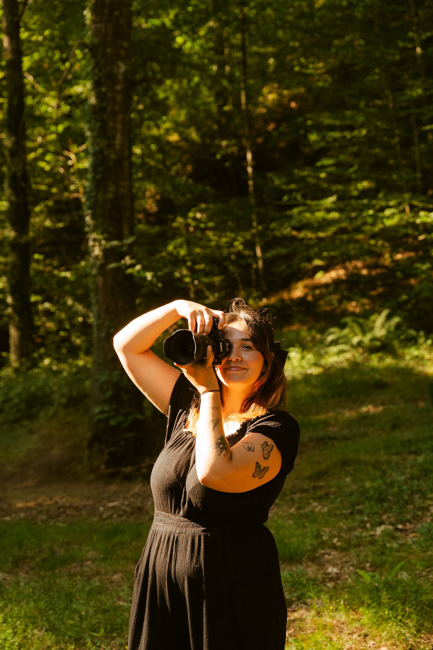 A photographer in a black dress takes photos in a sunlit forest setting.