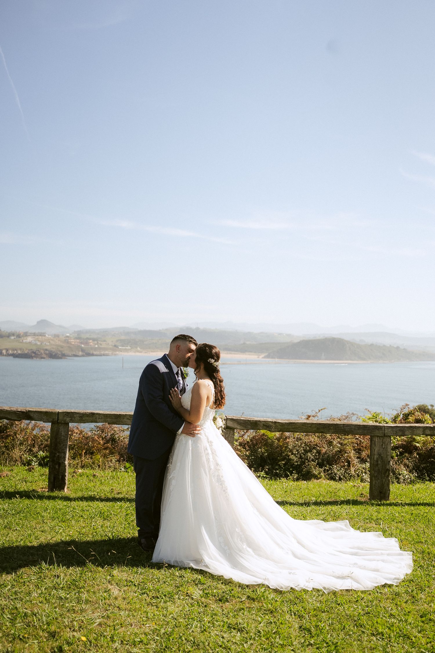 A couple in wedding attire shares a romantic moment on a hilltop overlooking a scenic coastal landscape.