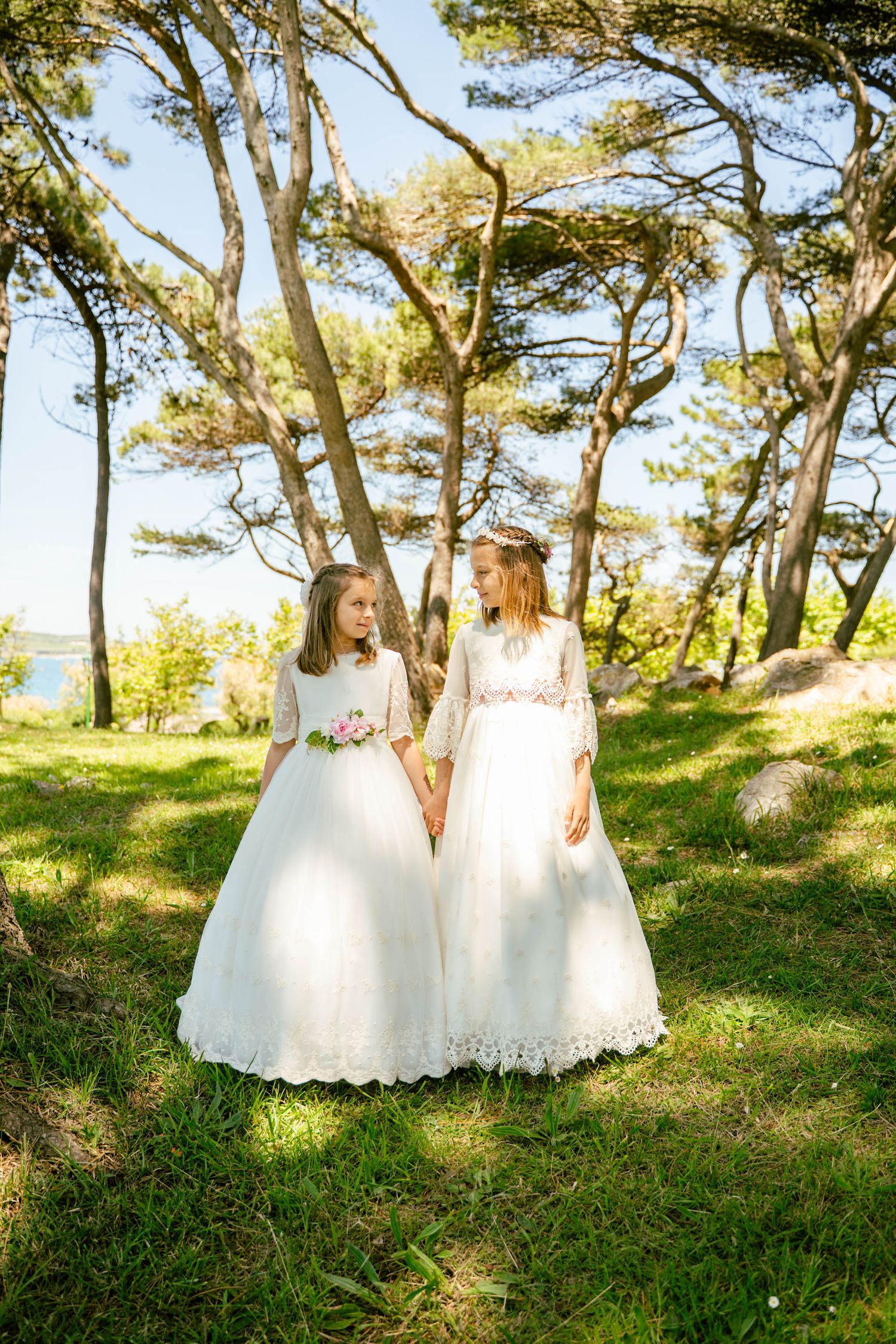Two white dresses photographed in a sunny coastal pine forest setting.