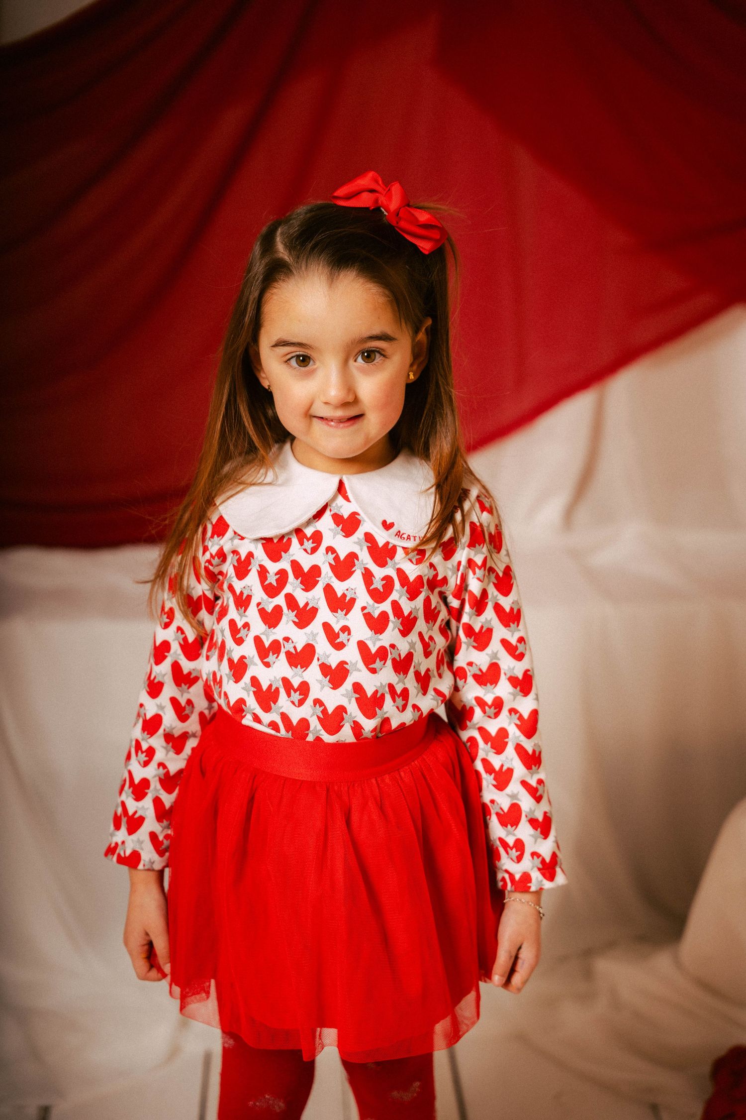 A child wearing a festive red and white heart-patterned dress with tulle skirt poses against a red and white backdrop.