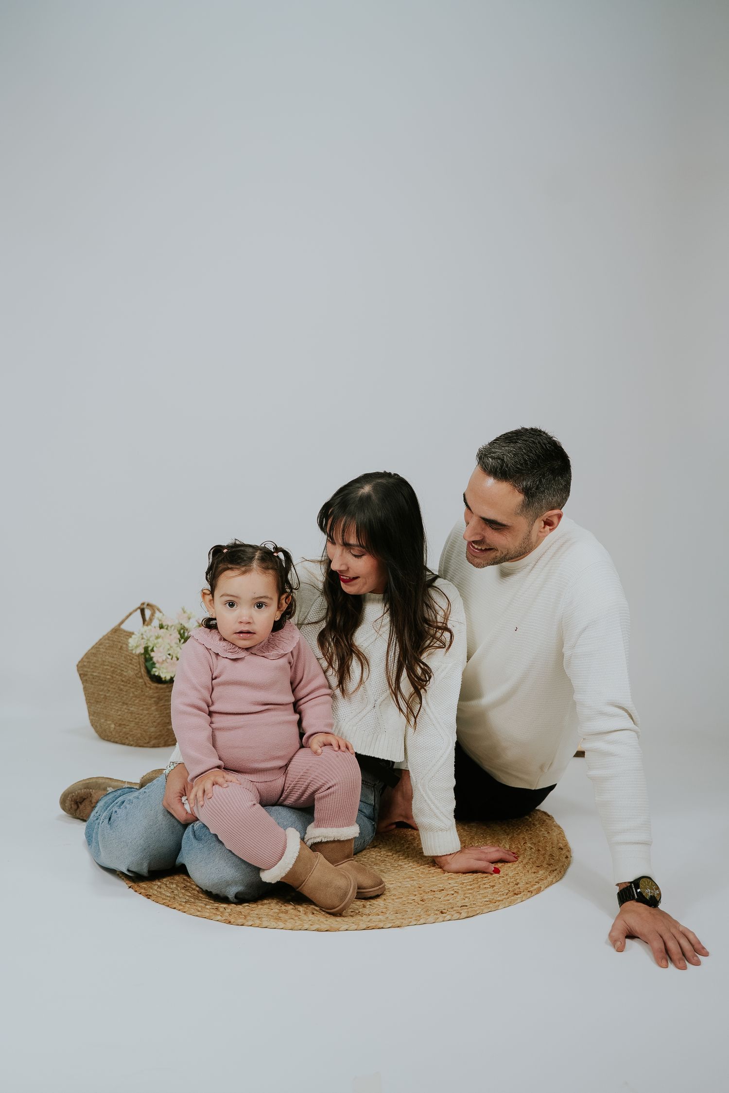 A family sits together on a round jute rug in a minimalist setting wearing coordinated white and pink outfits.