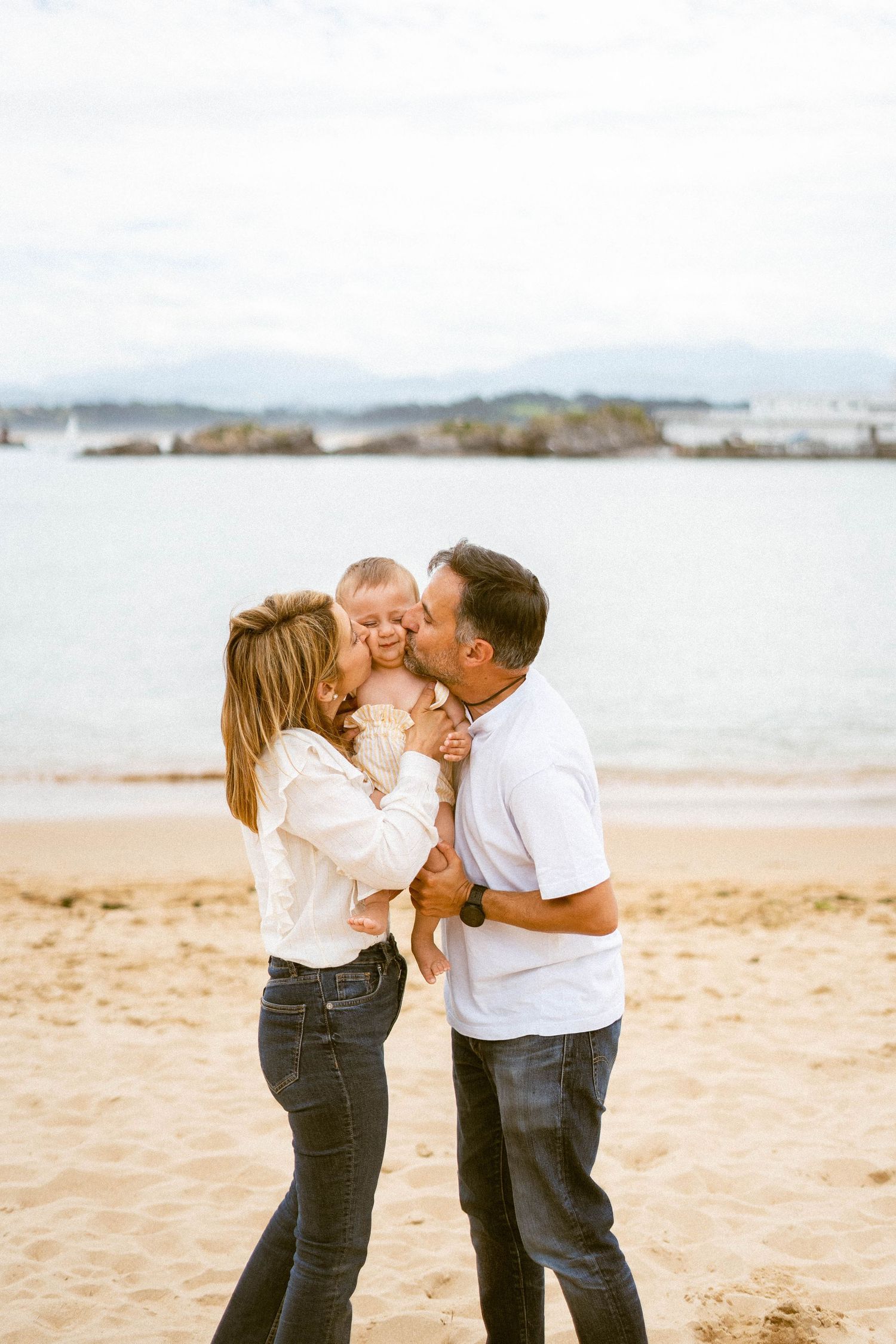 Couple shares tender moment on sandy beach in casual white and denim outfits.