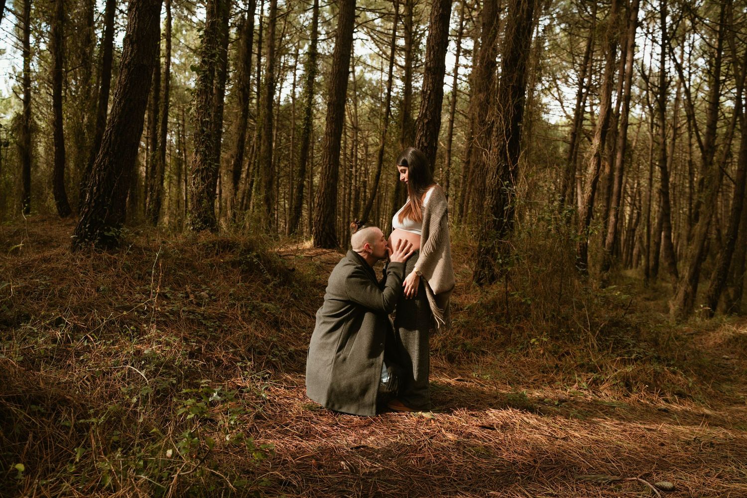Tender pregnancy moment captured in a forest setting with pine trees in the background.