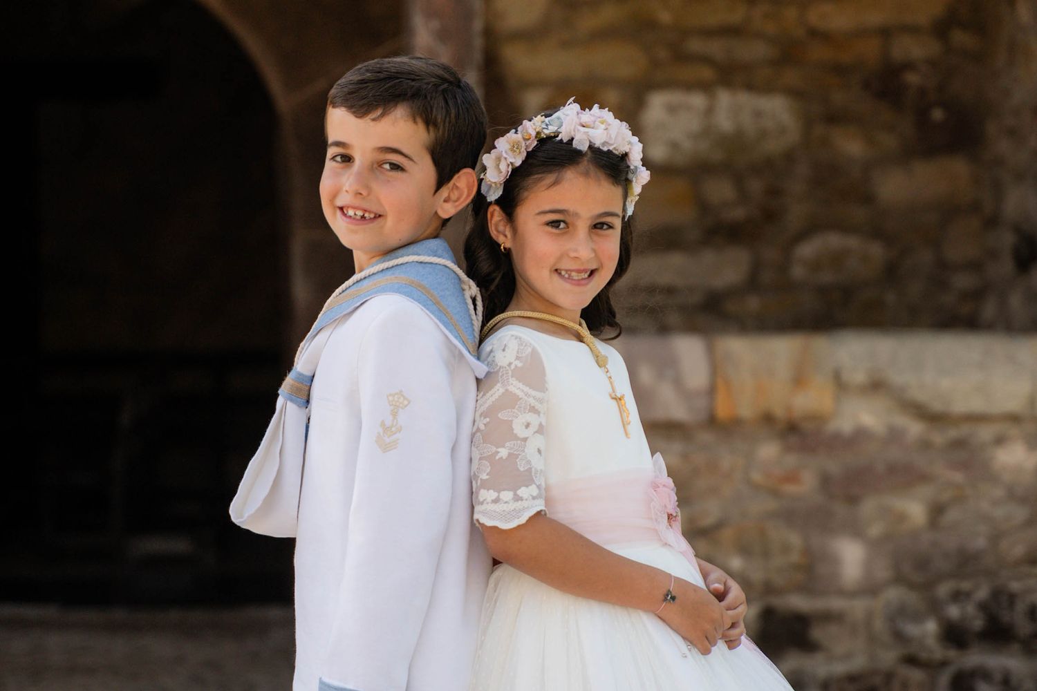 Children in white formal attire pose together against rustic stone wall background.