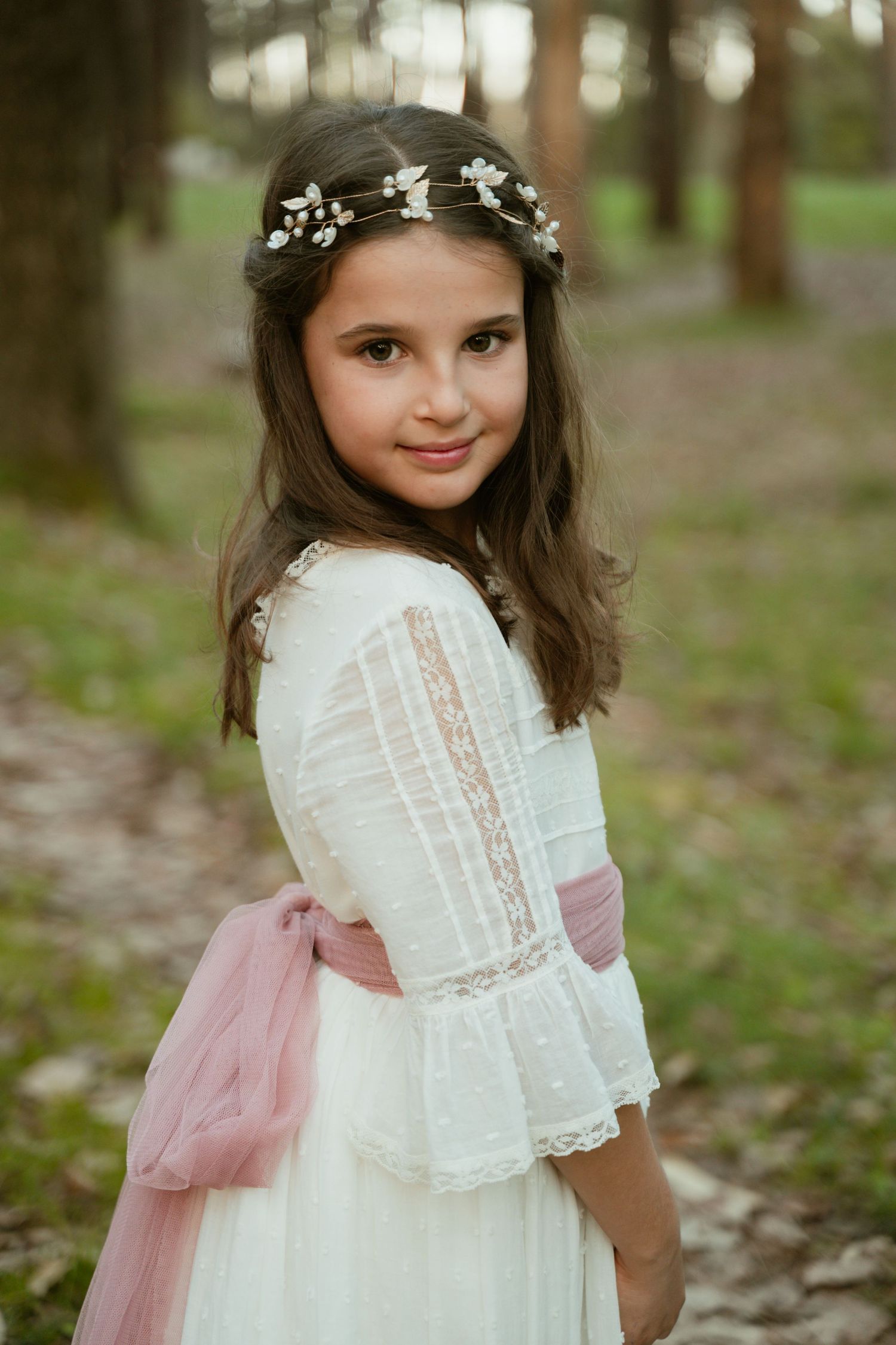 Delicate lace dress with pink sash and floral crown captured in a wooded setting.
