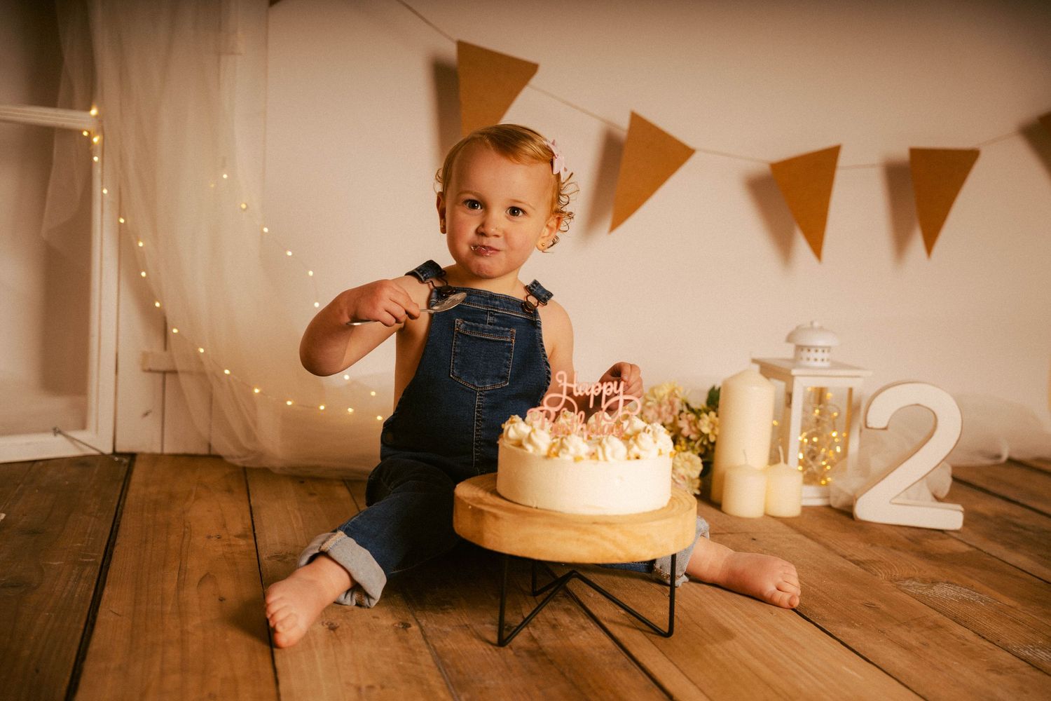 A child in denim overalls sits on a wooden floor with fairy lights and decorative banners in the background.
