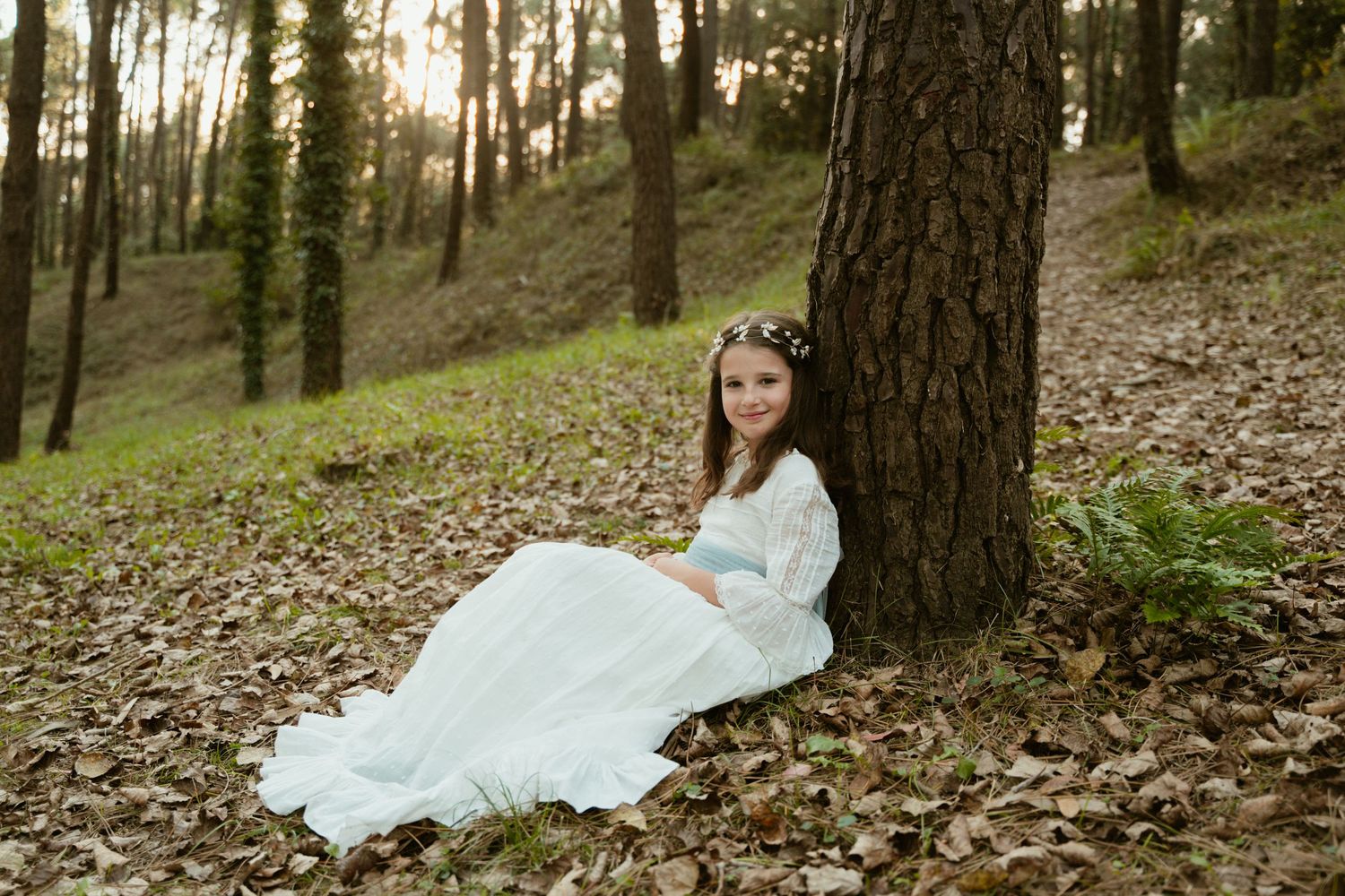 Whimsical forest scene with white dress and crown while sitting against a pine tree.