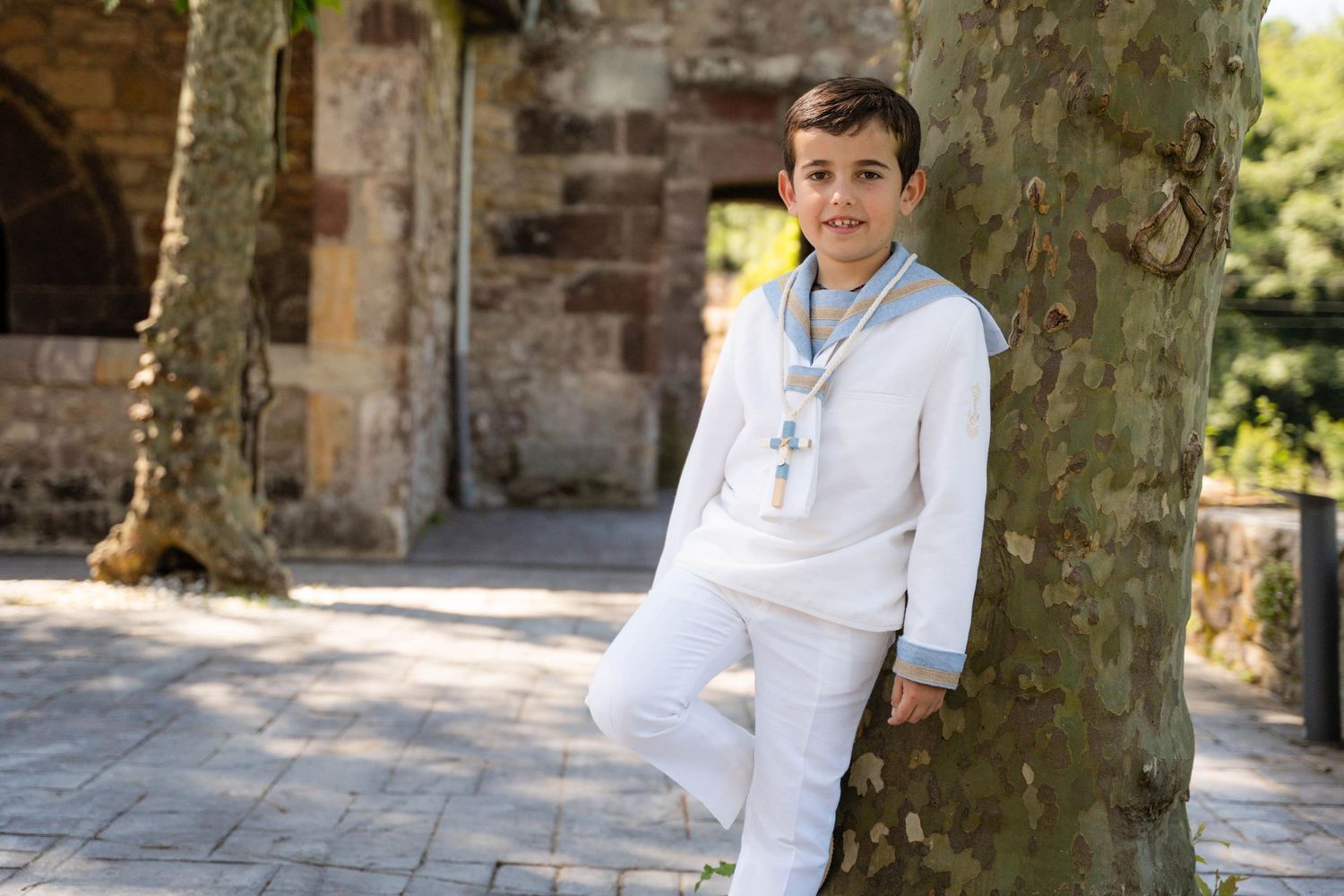 Children in white formal attire pose against stone building and trees on sunny day.