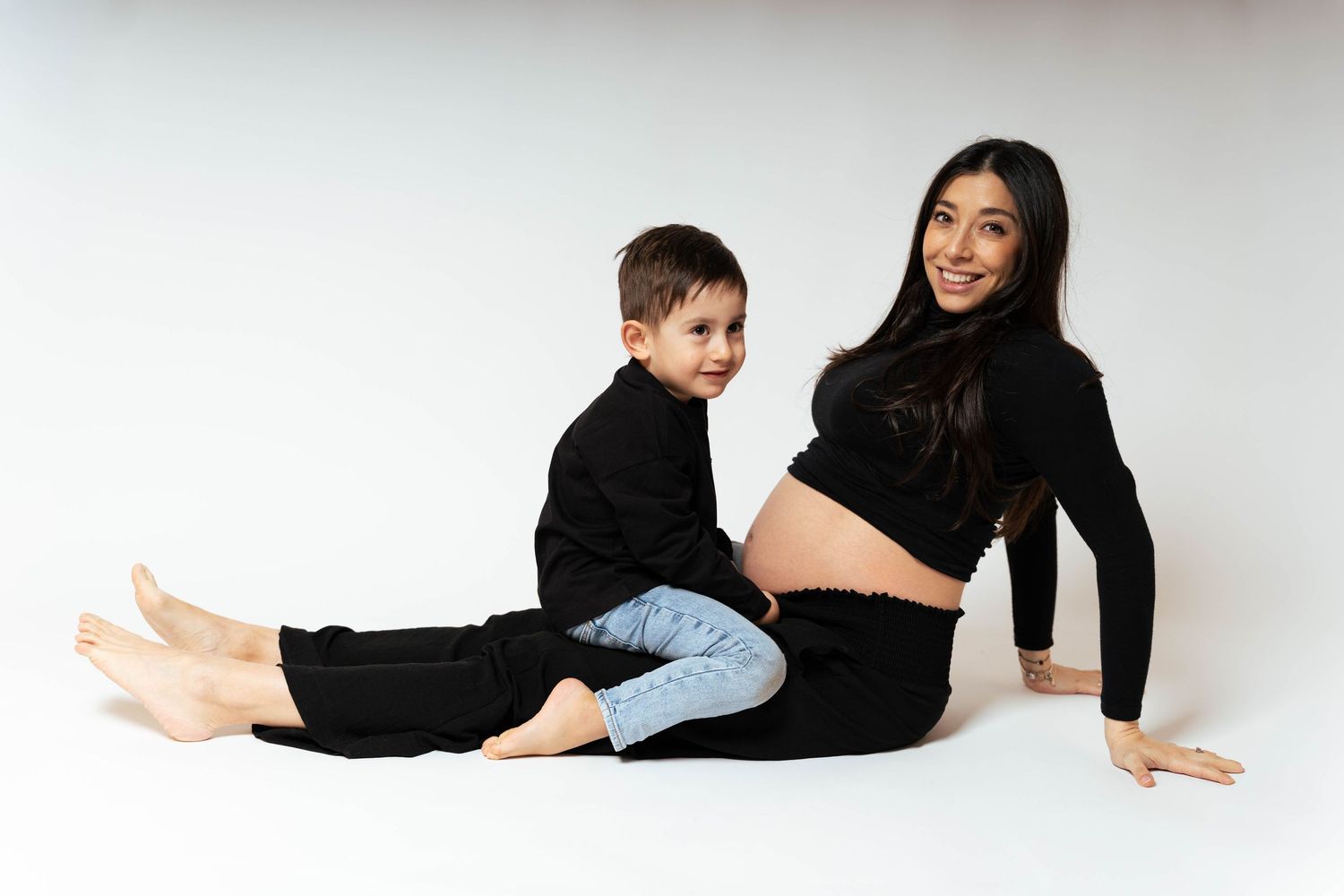 A maternity photo on white background shows a woman in black sweater with child in casual attire sitting together.