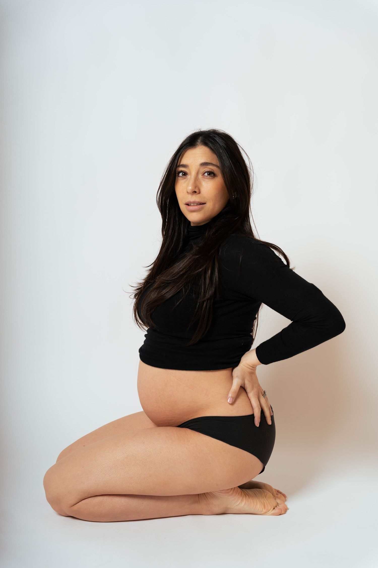 Minimalist maternity photo of woman in black long sleeve top and shorts kneeling against white background.