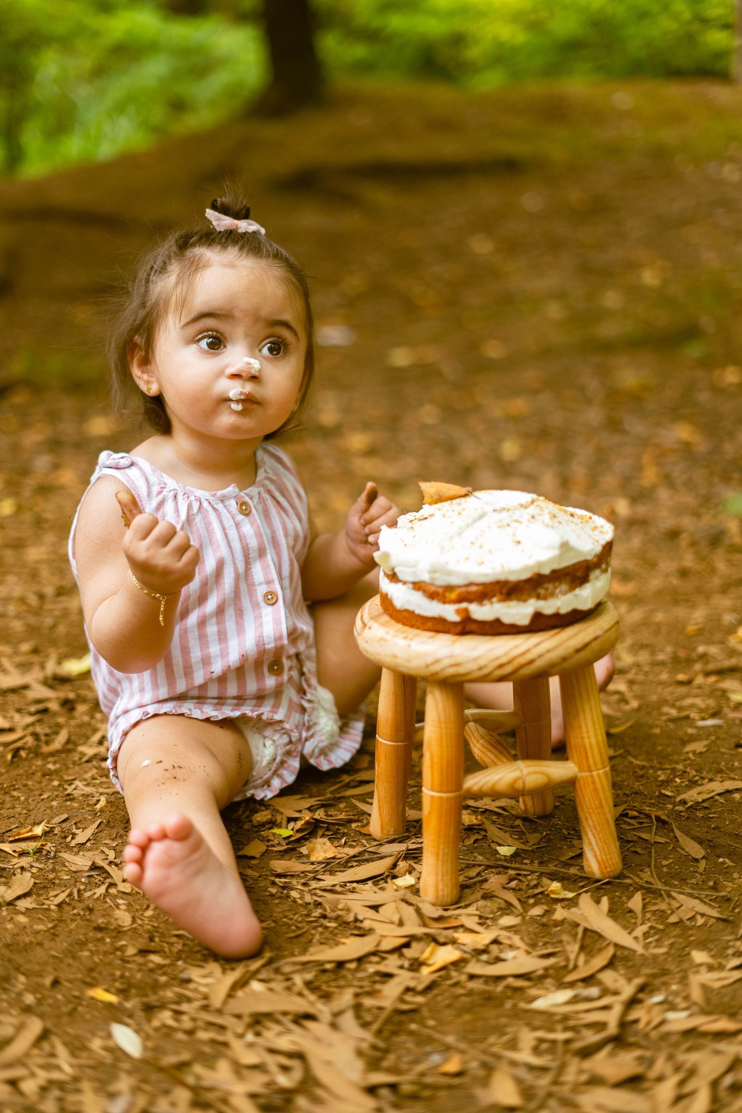 A small wooden stool holds a cream-filled cake on a forest floor with scattered leaves.