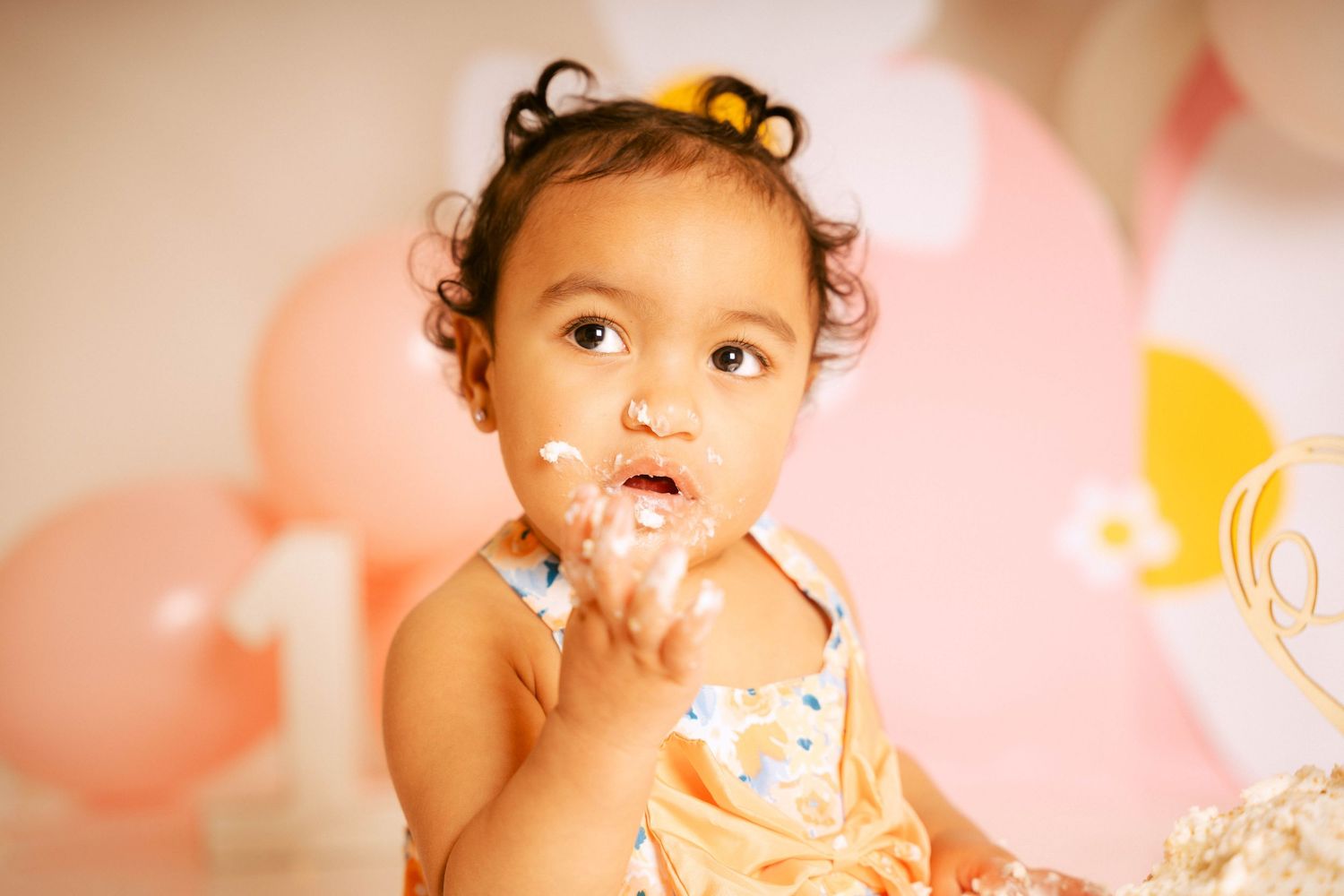 A toddler in a pink dress celebrates their first birthday with cake smash photos against a pink and white backdrop.