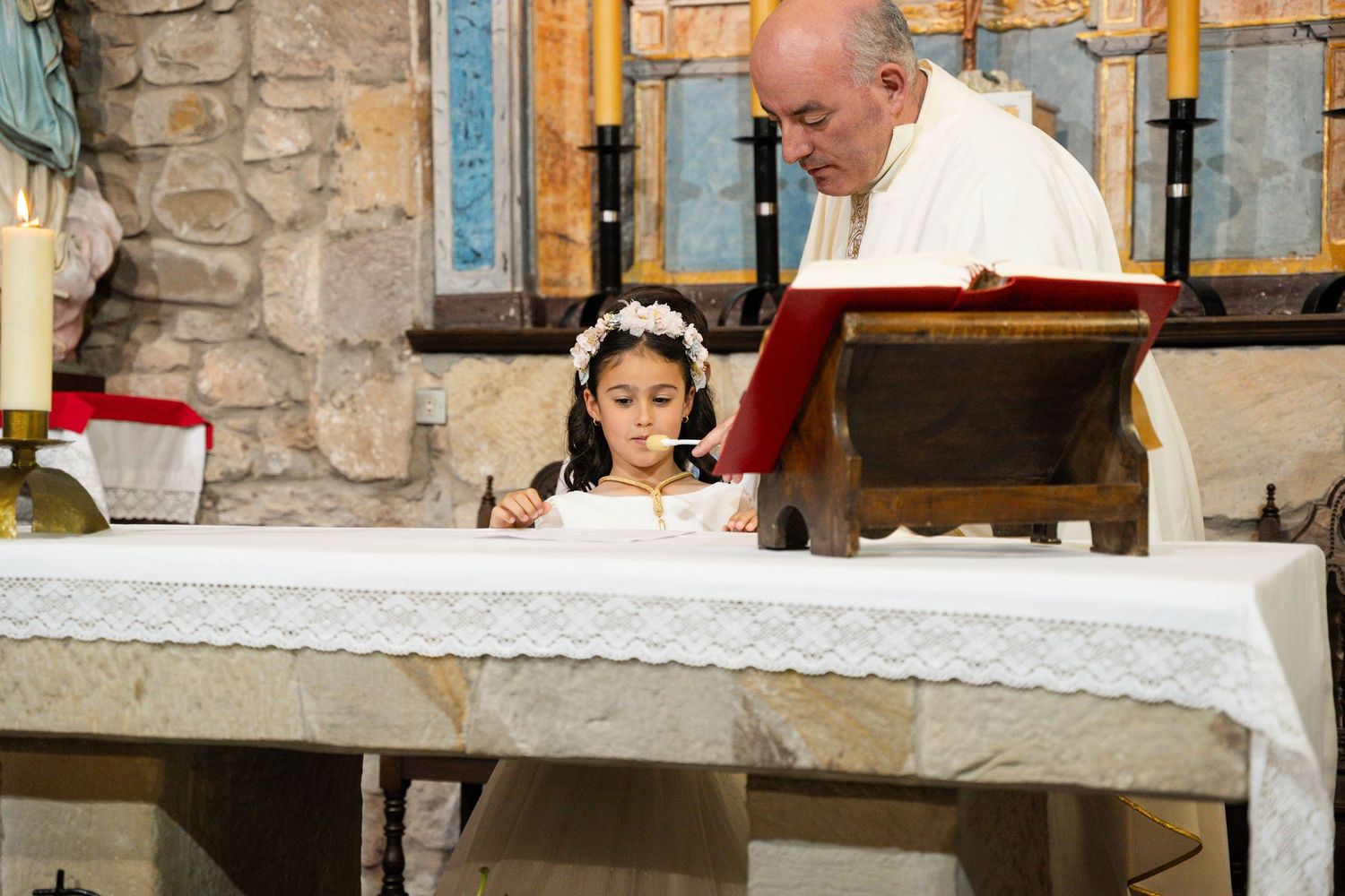 A young child in white attends a religious ceremony at a stone church altar.