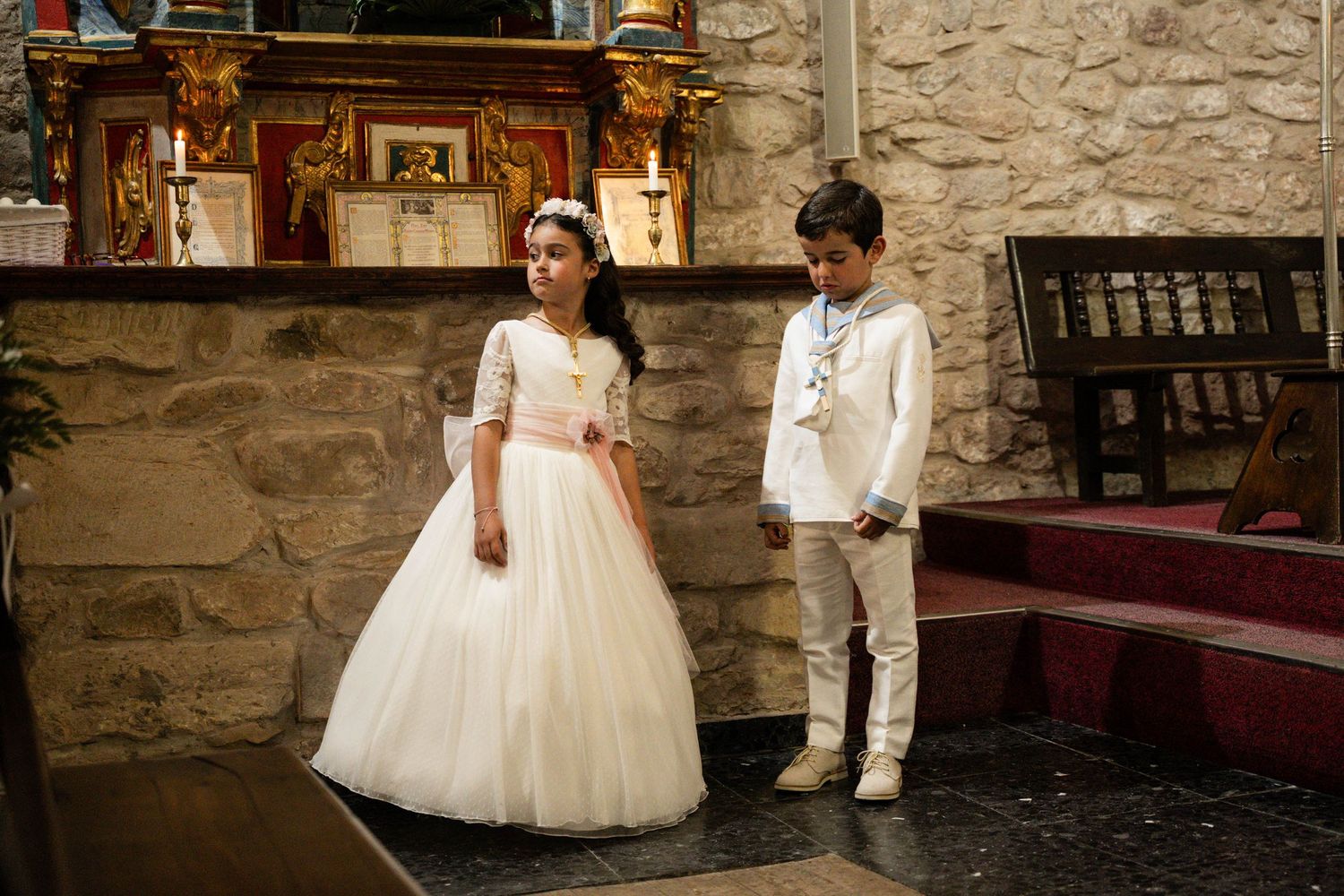 Children in formal white outfits stand inside historic stone church with religious artwork.