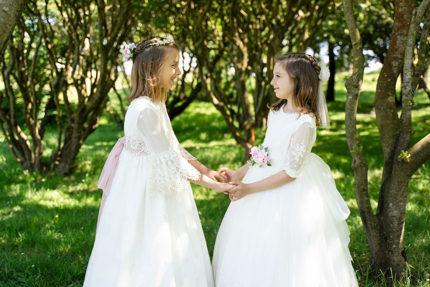 Two children in white formal attire pose together in a sunny garden setting with lush green trees in the background.