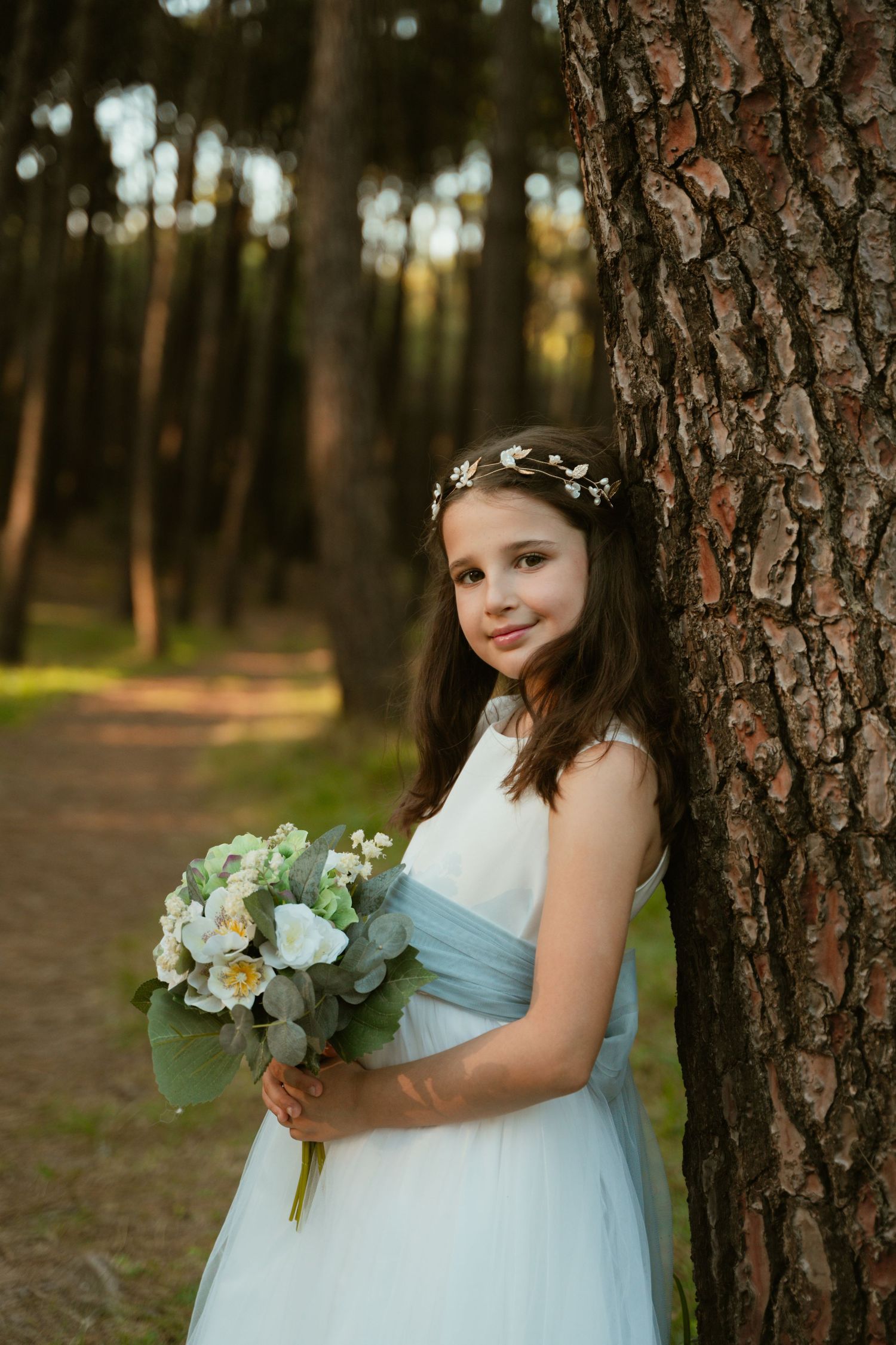 Young bridesmaid in light blue dress holds eucalyptus bouquet against pine trees.