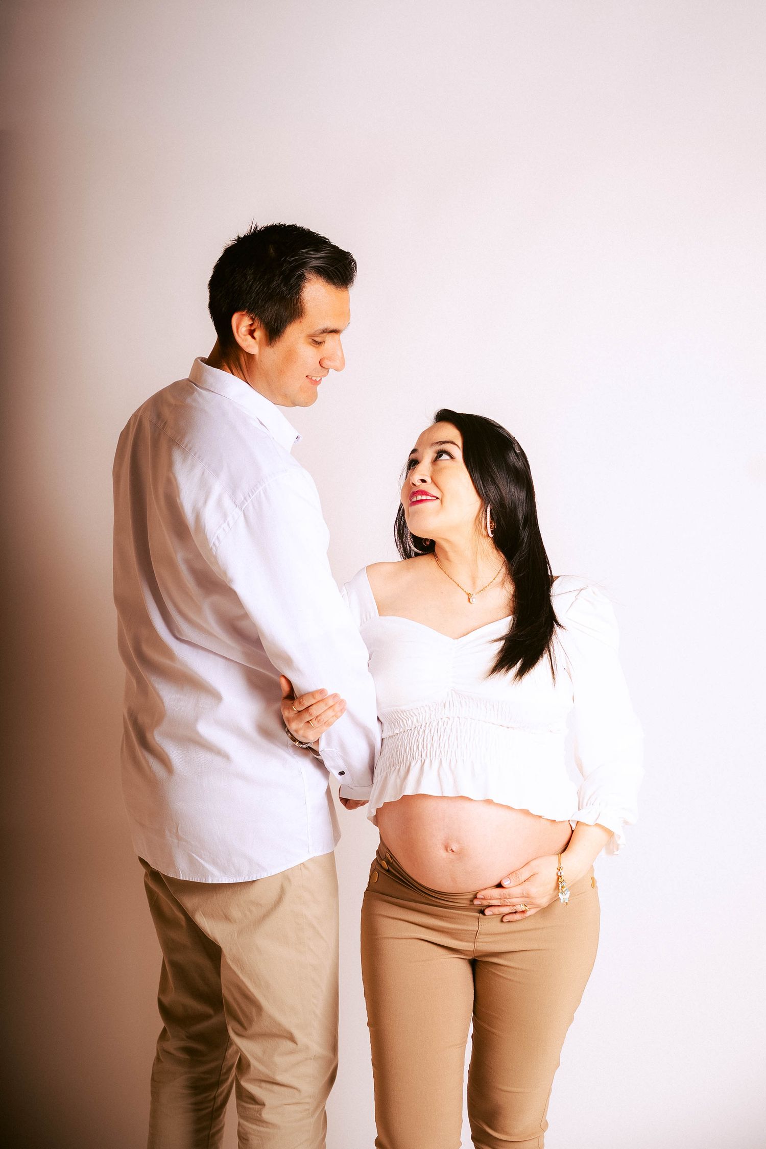 A couple in white and khaki outfits share tender moments during a maternity photoshoot against a light backdrop.