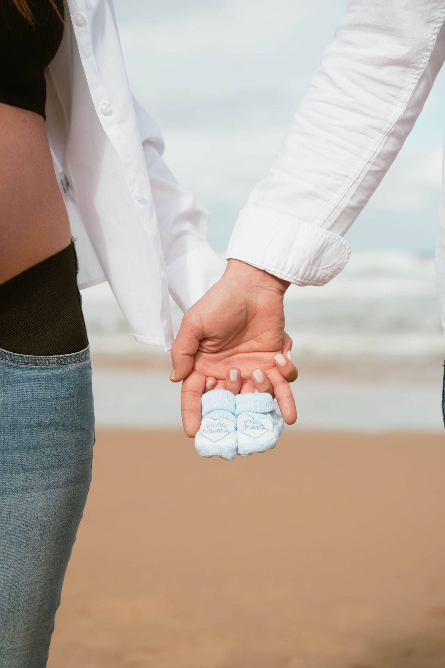 Couple holding tiny baby shoes together on a beach, symbolizing their upcoming parenthood journey.