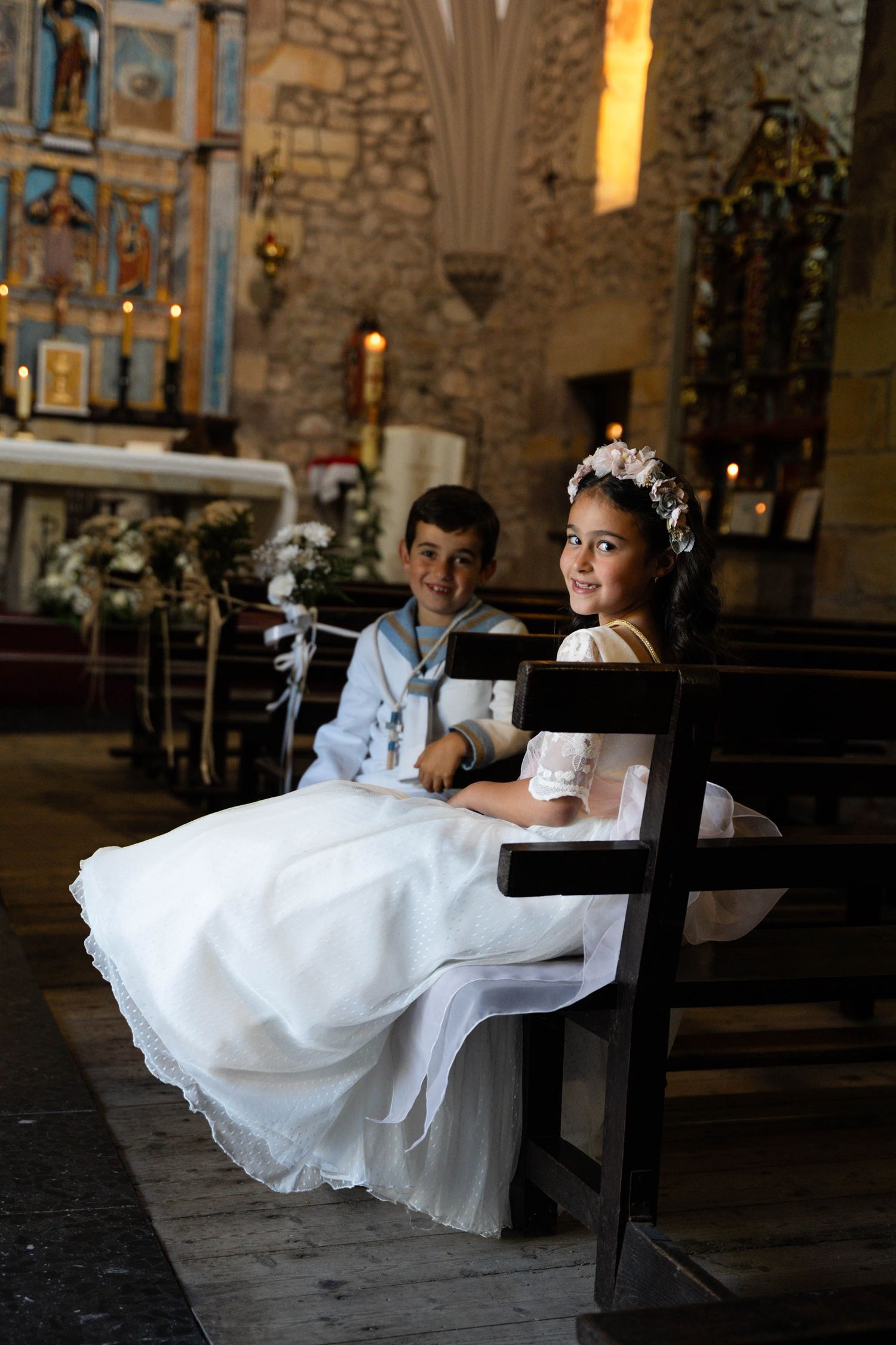 Children in formal white attire sit on wooden church pews inside historic stone building.