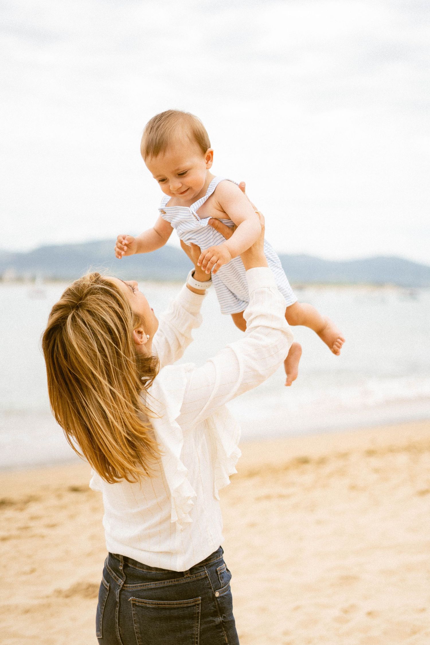Parent playfully lifts baby in white outfit on sunny beach.