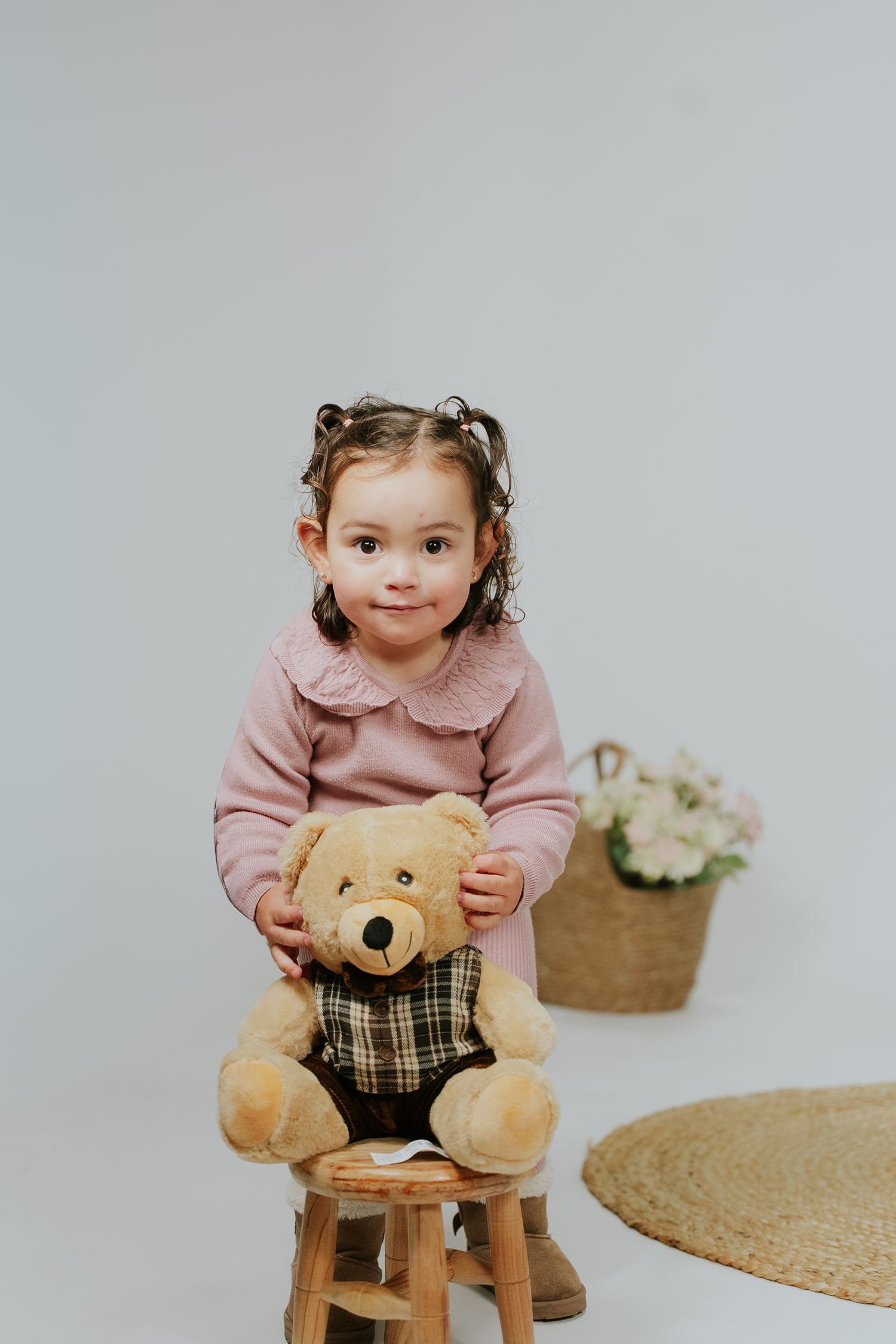 A small child in a pink sweater sits on a wooden stool with a plush teddy bear wearing plaid clothing.