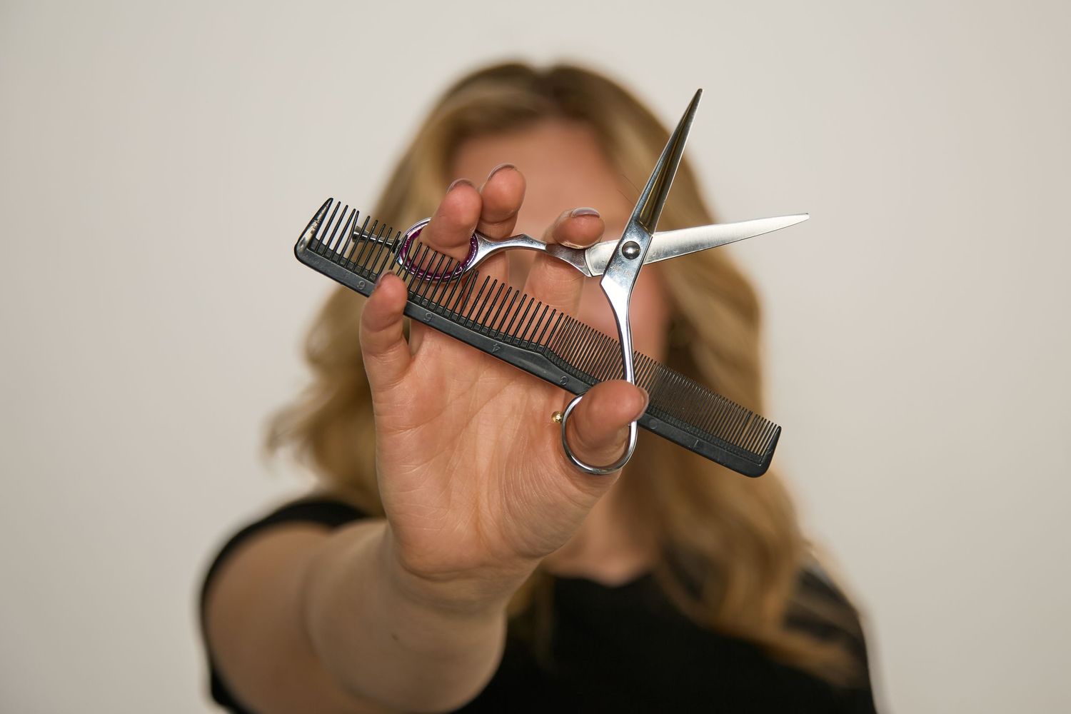 Blonde woman holding hair scissors and comb toward camera in a professional hairstylist pose.
