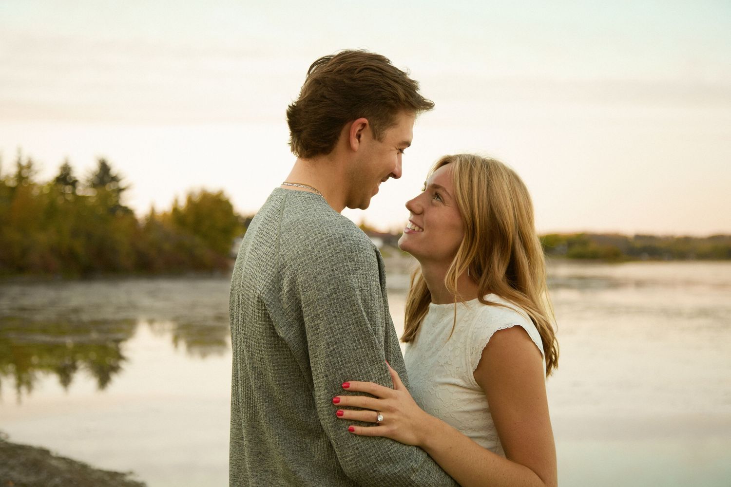 A young couple shares a romantic moment by a lakeside at sunset, smiling at each other tenderly.