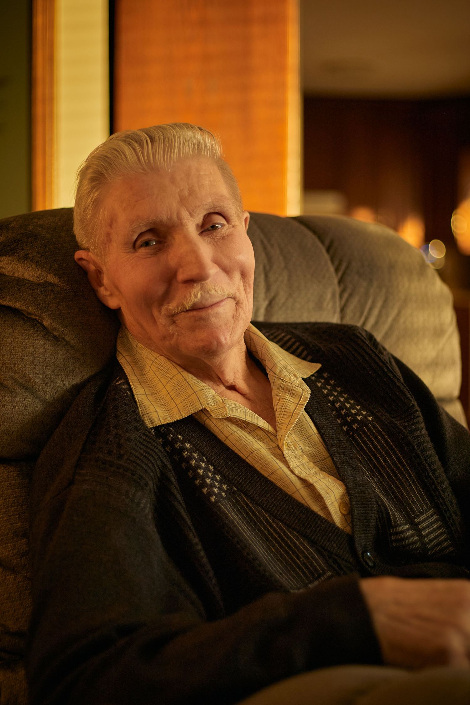 An elderly person in a dark sweater rests on a couch in warm ambient lighting.
