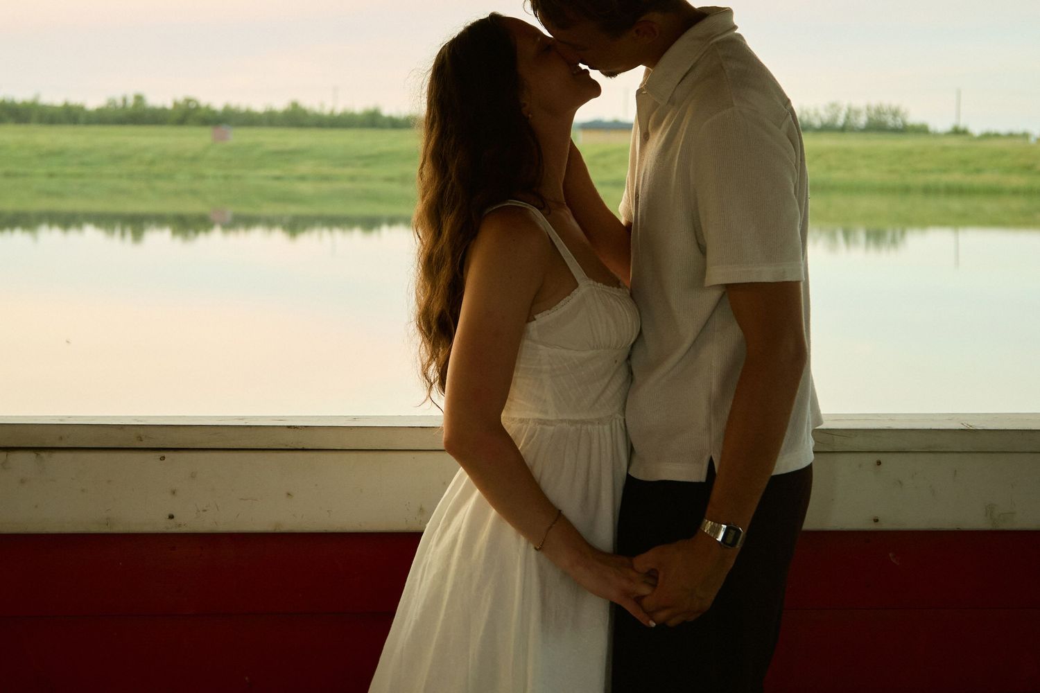 Romantic couple sharing a kiss in white outfits by a serene lakeside window at sunset.