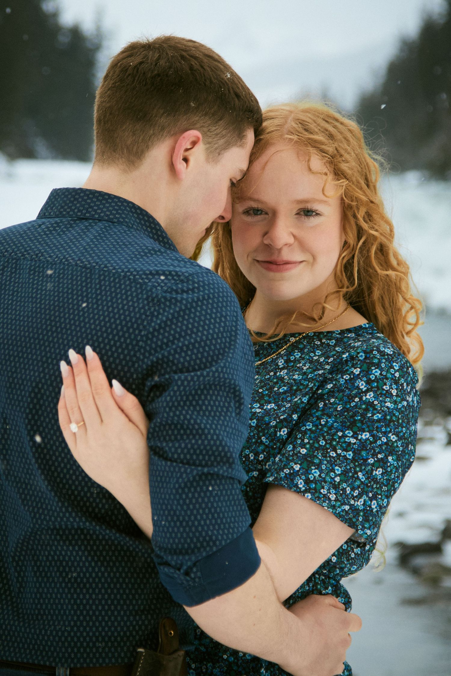 Couple embracing in snowy winter landscape, woman with curly red hair smiling, wearing floral dress, engagement ring visible.