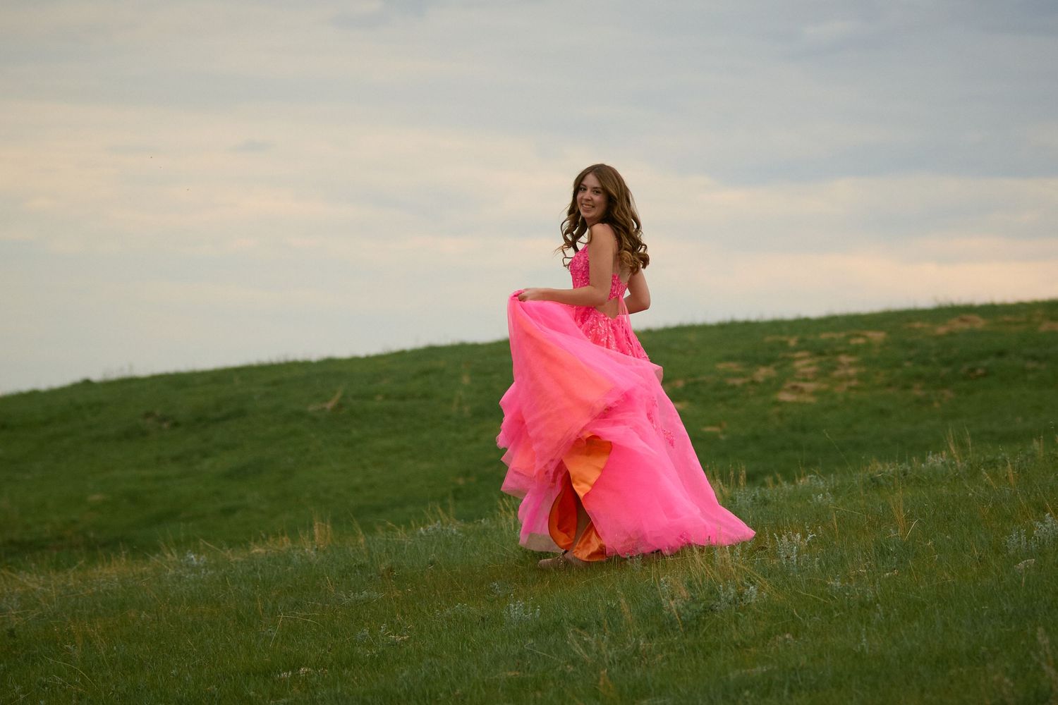 Young woman in a flowing pink ball gown twirling on a green hillside at sunset.
