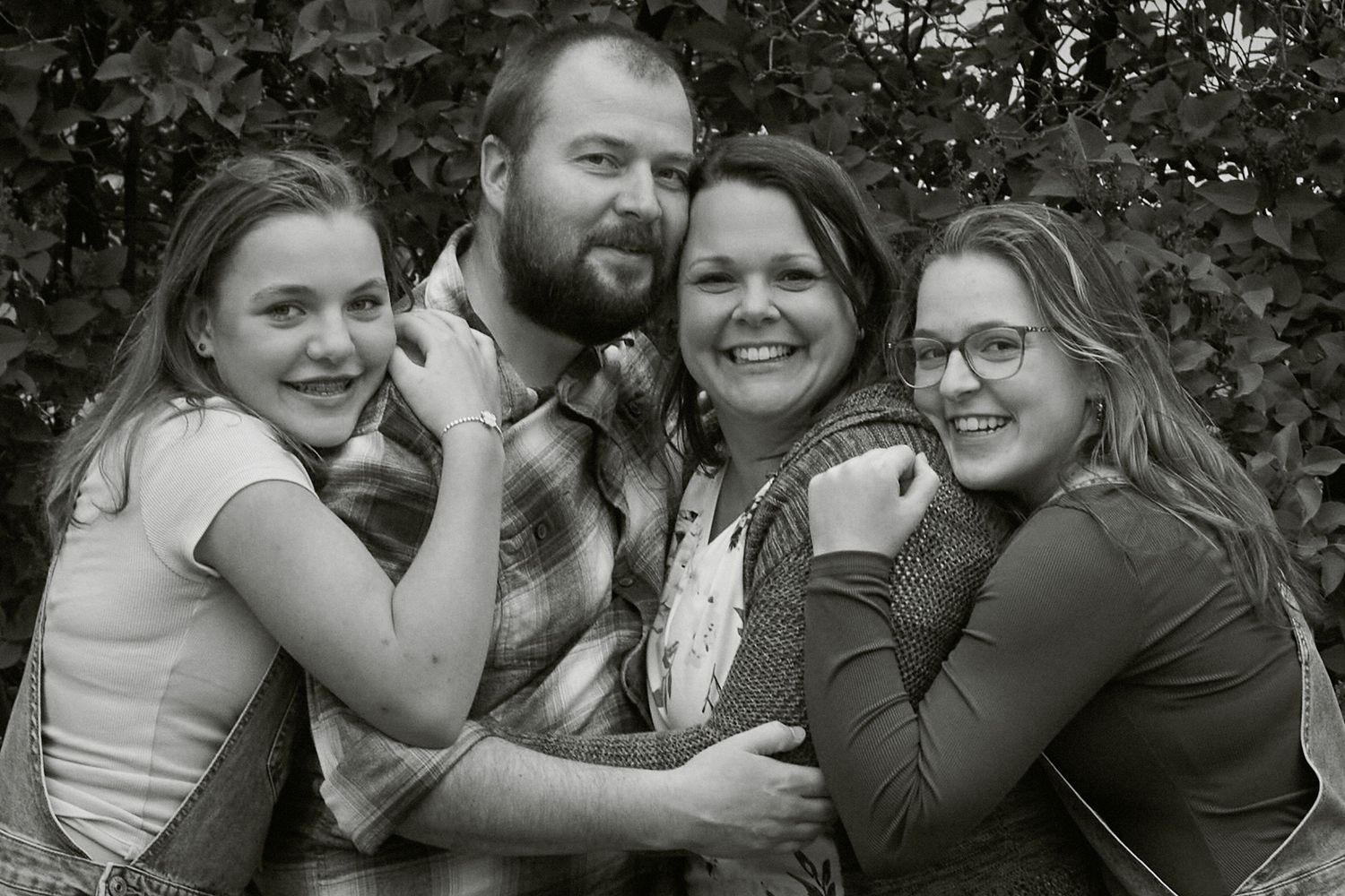 Black and white family portrait of smiling parents and two teenage daughters embracing outdoors.