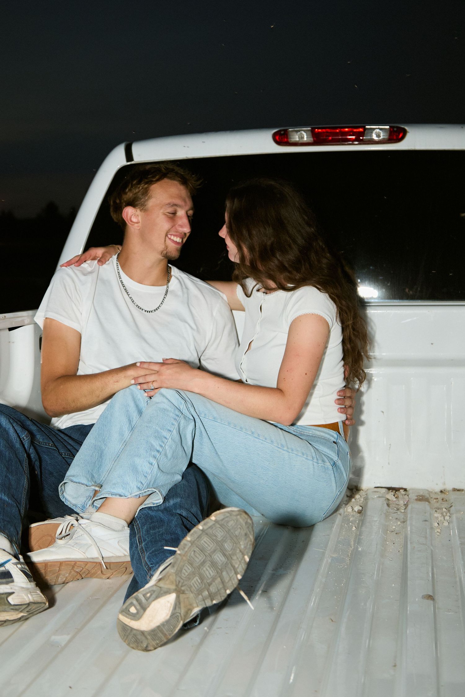 A smiling young couple sits closely together in the bed of a white pickup truck under a night sky.