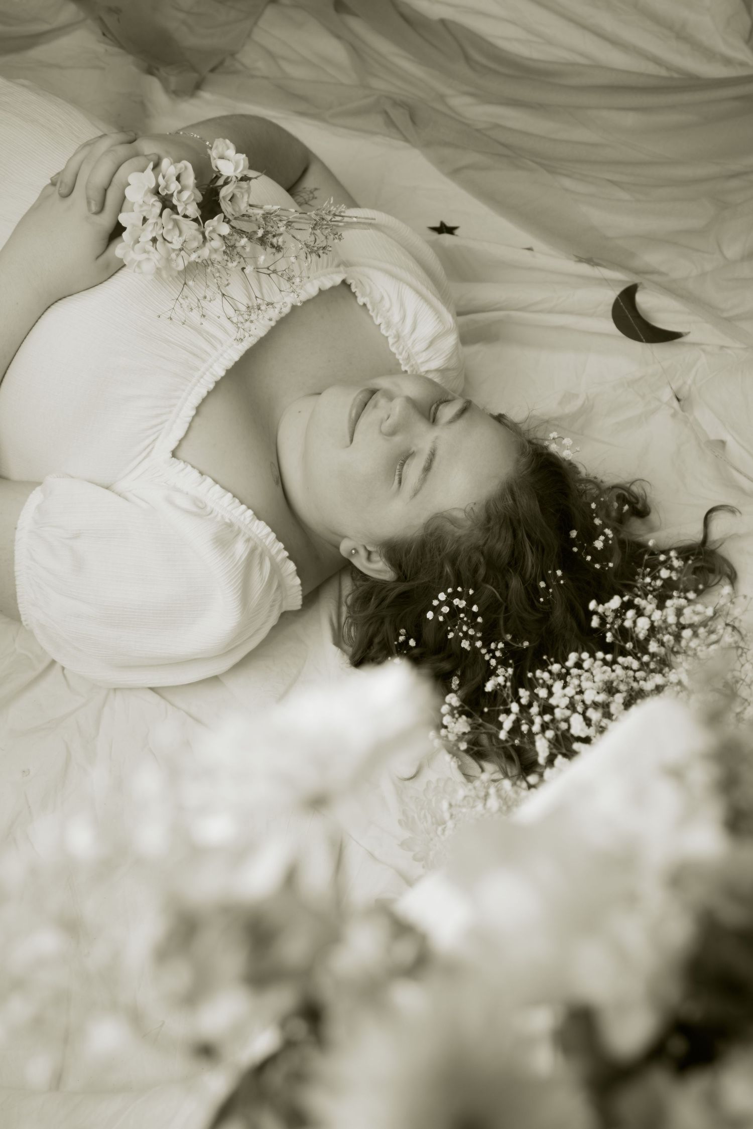 Dreamy black and white portrait of someone lying on fabric with delicate white flowers in their hair and dress.