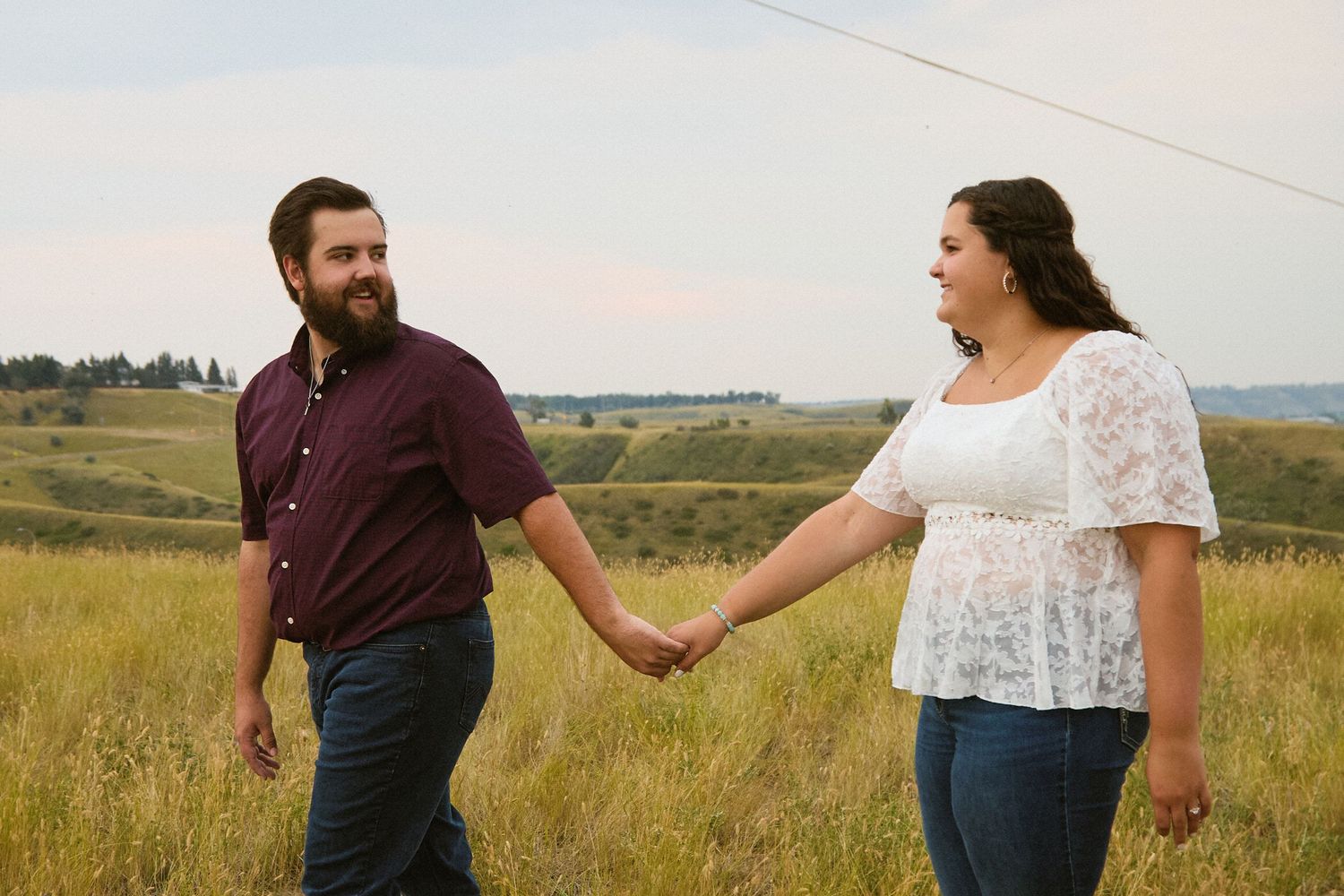 Couple holding hands in a grassy field with rolling hills, man in maroon shirt and woman in white lace top.